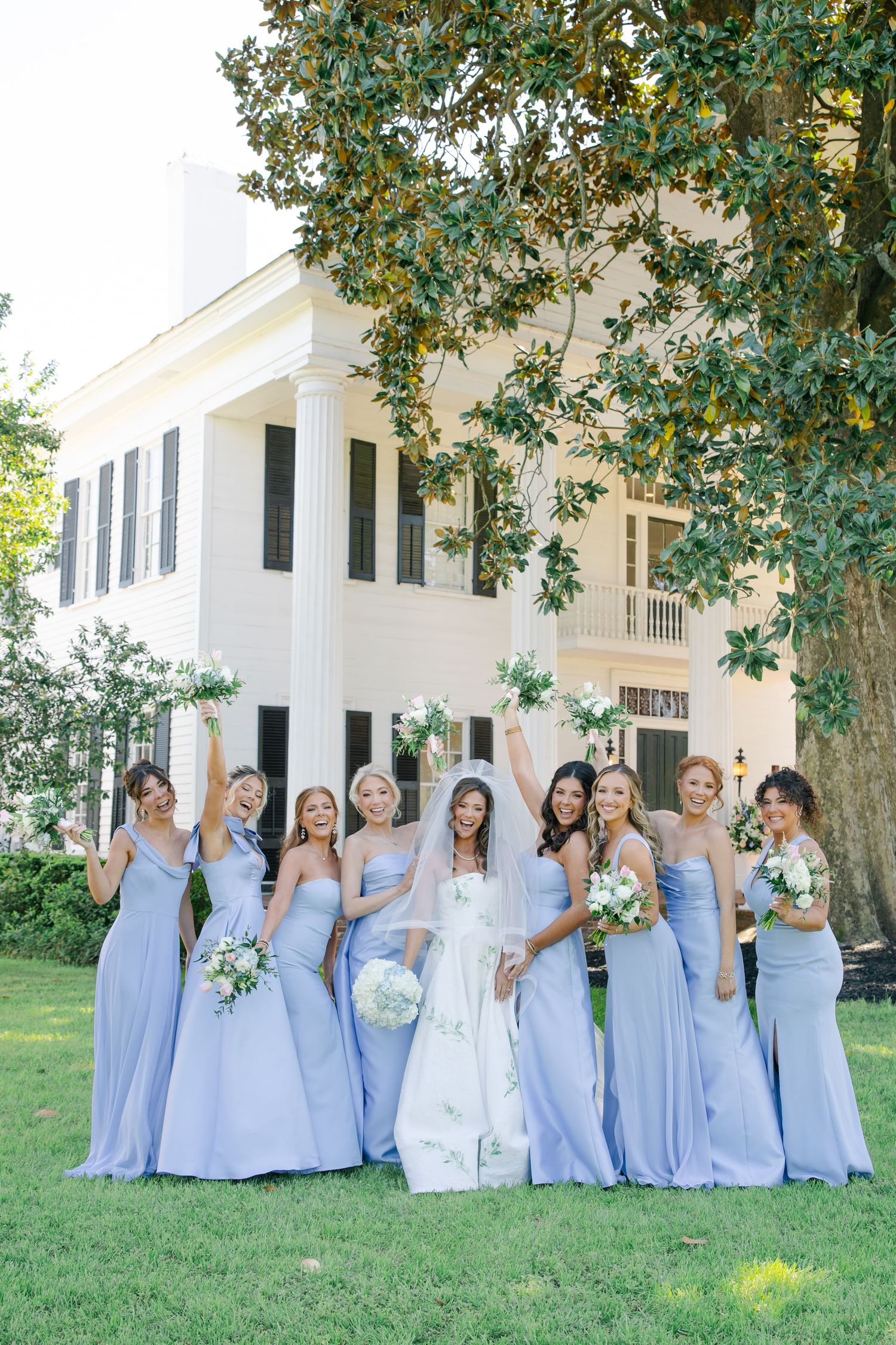 Joyful bridal party raises bouquets in celebration wearing periwinkle gowns outside a grand white Southern plantation home.