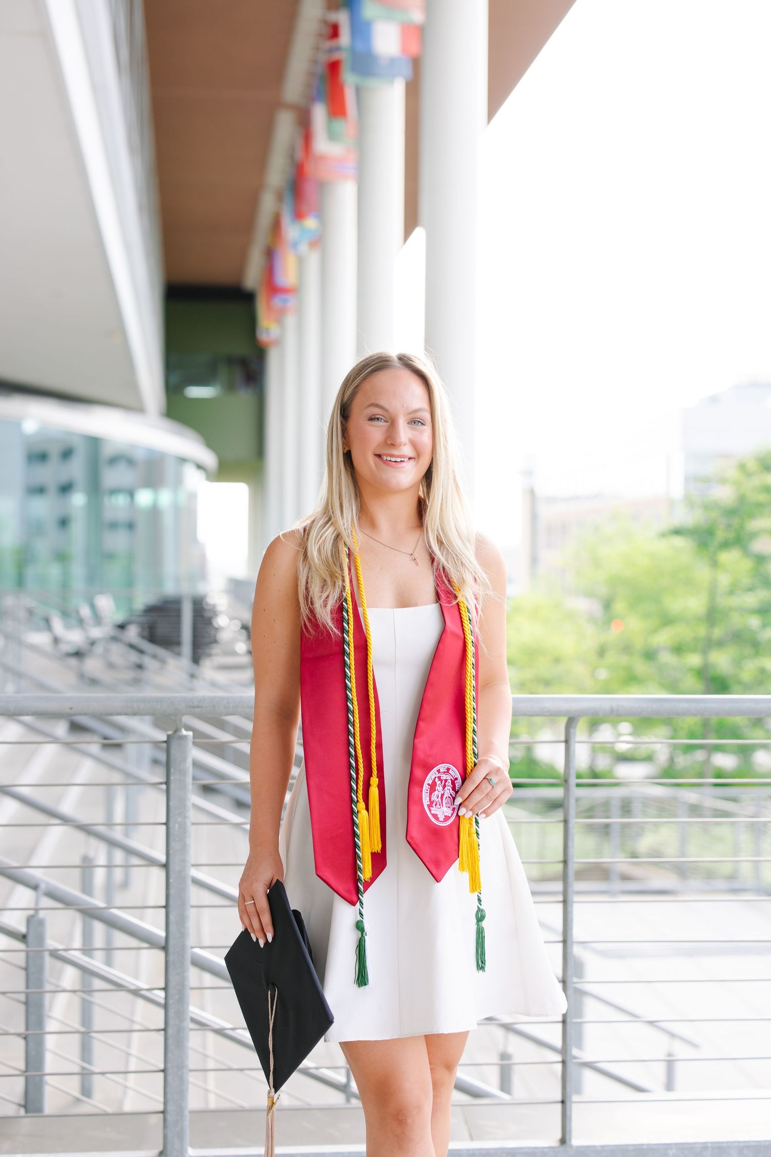 Smiling blonde graduate in white dress with honor stoles holding cap at university.