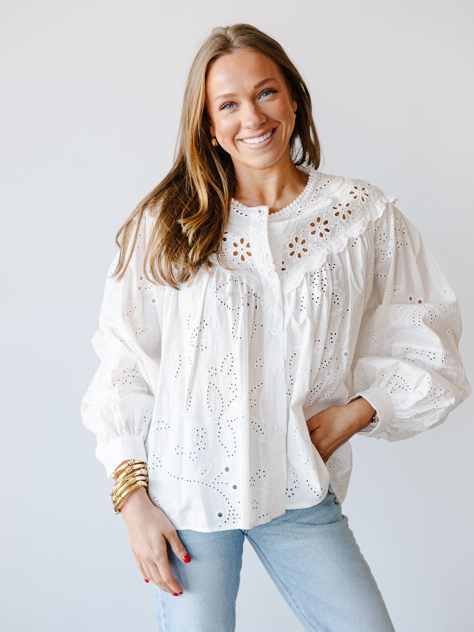 Smiling woman wearing a white eyelet lace long sleeve blouse with gold bangles and blue jeans.