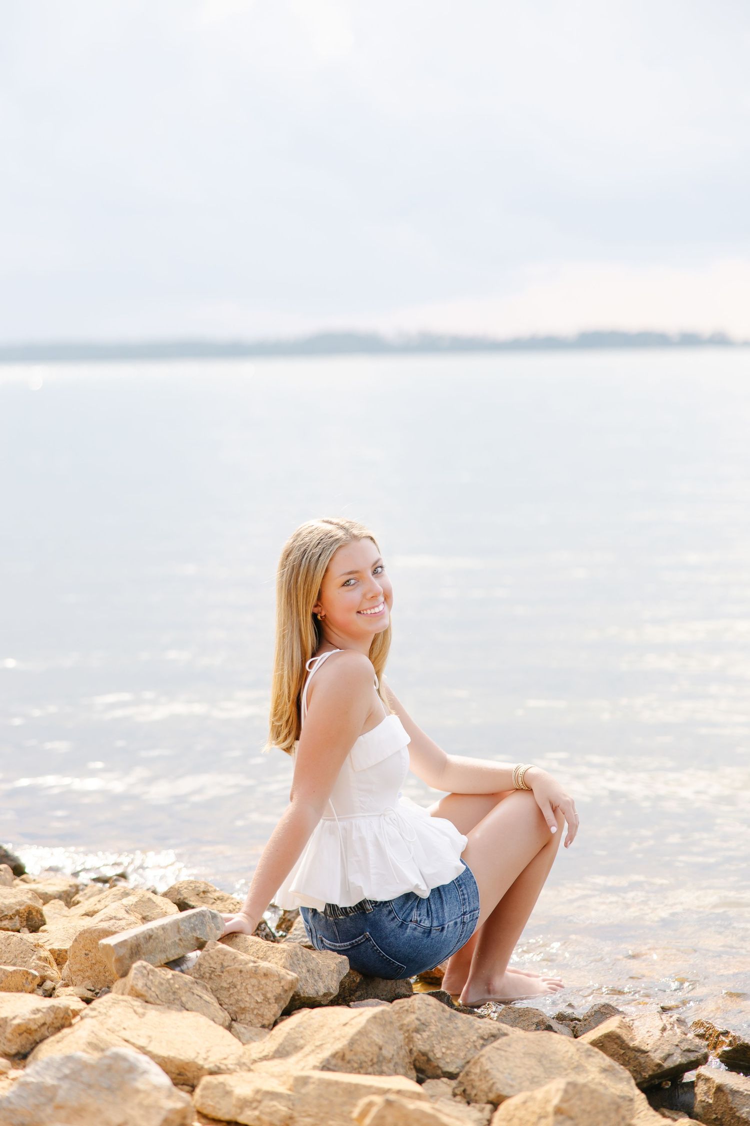 Smiling blonde teen in white top and denim shorts sitting on rocks at waters edge by lake.