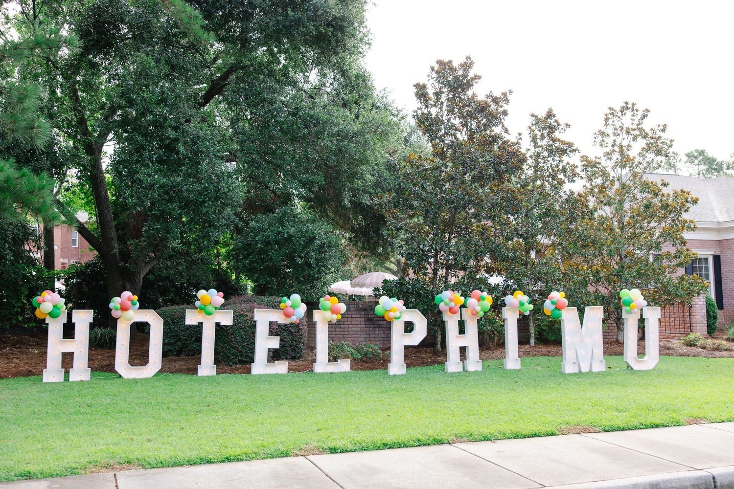 Large white marquee letters spelling HOTEL PHI MU decorated with colorful balloons on a green lawn.