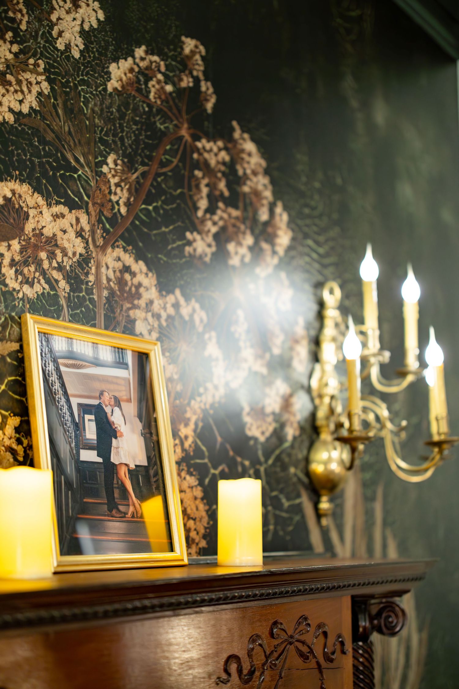 Gold framed couple photo on antique mantel with glowing candles and brass candelabra against dark floral wallpaper.