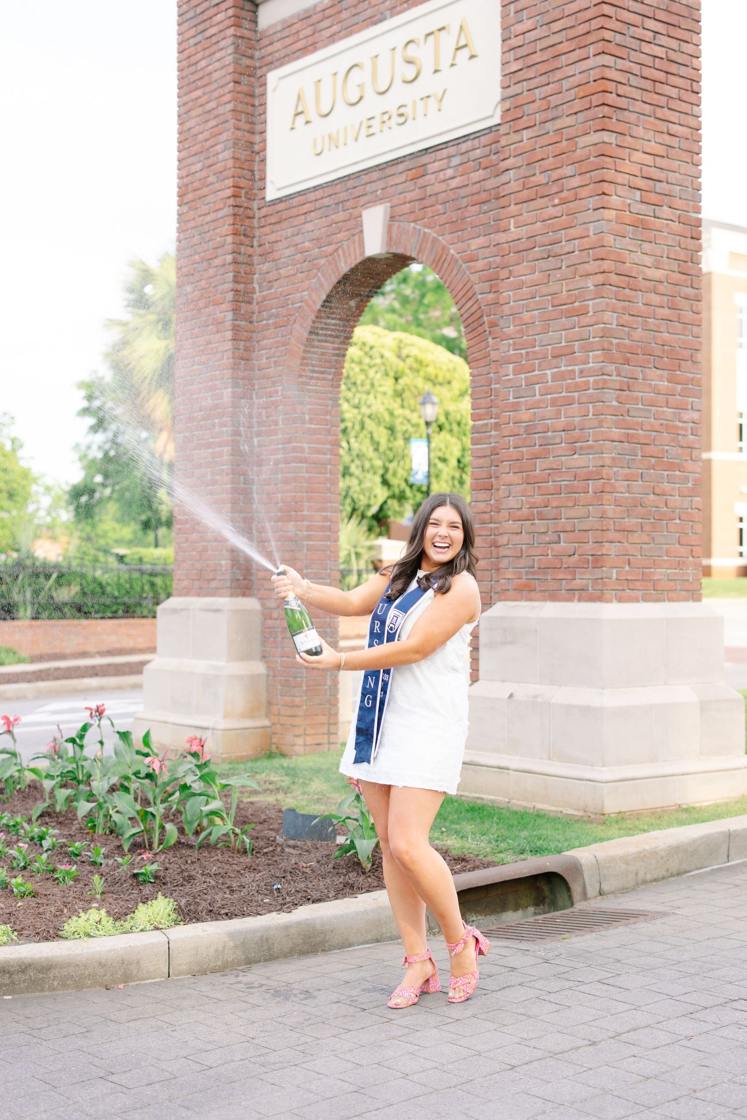 Excited Augusta University nursing graduate spraying champagne in front of brick university arch.