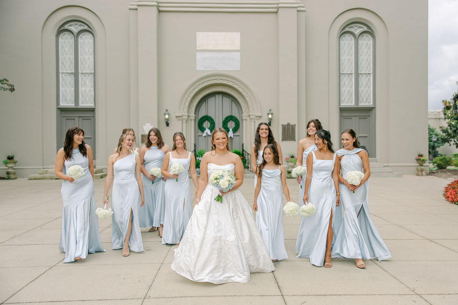 Bride in white ball gown walks with bridesmaids in light blue satin dresses holding white bouquets outside a elegant church.