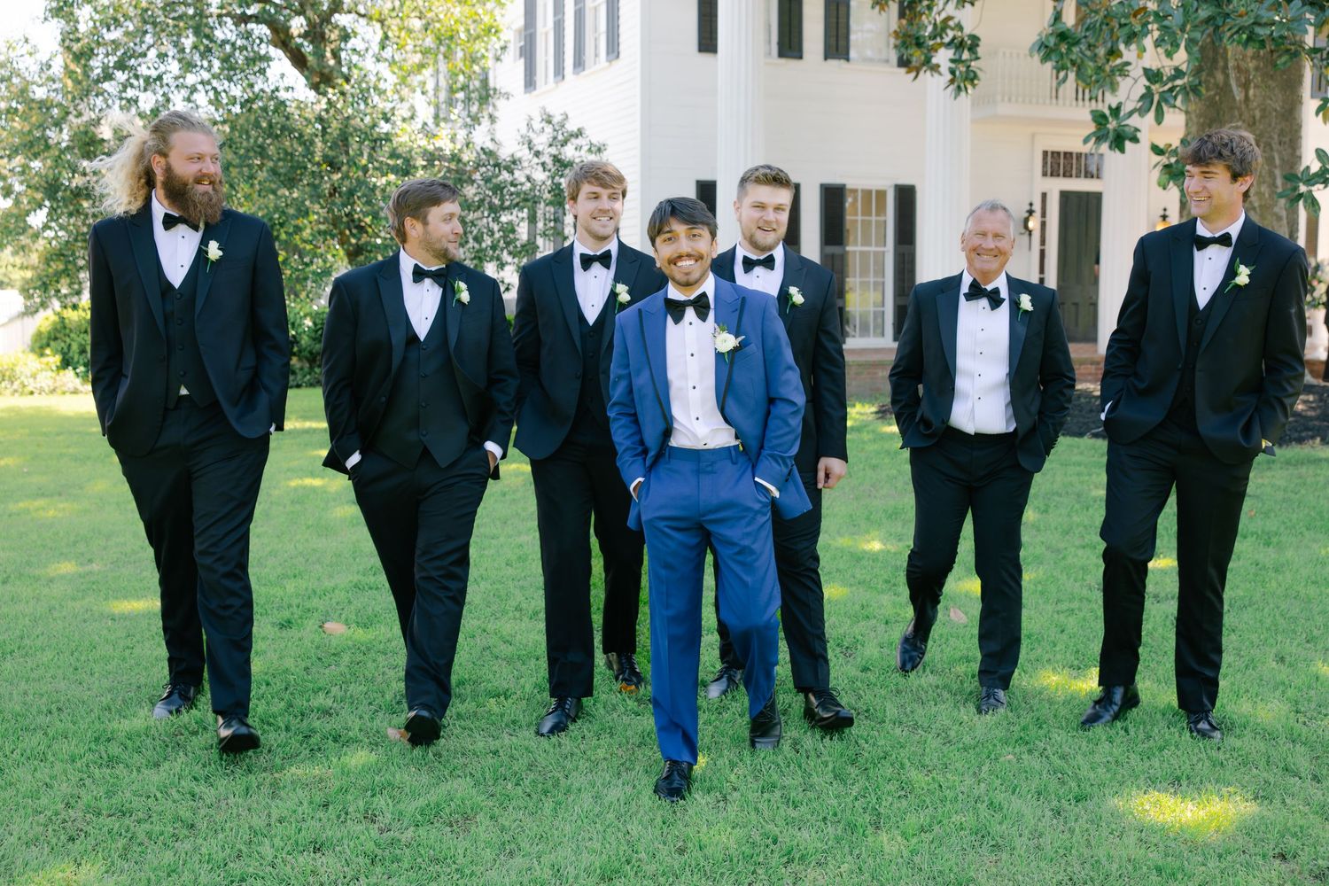 Groom in blue tuxedo walking with groomsmen in black tuxedos on lawn.