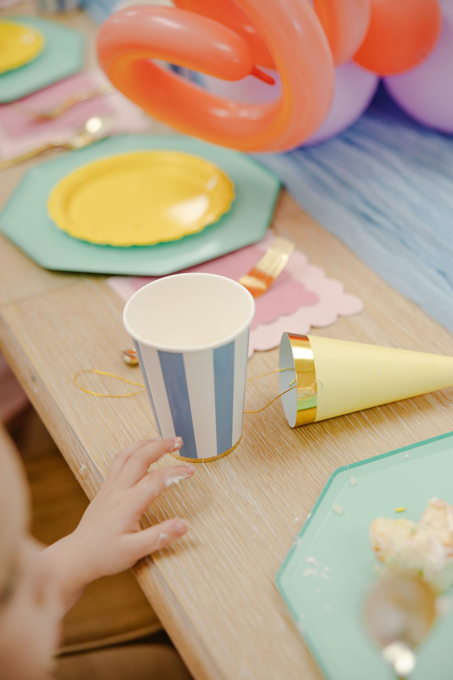 Child's hand reaching for a blue striped paper cup near a yellow party hat on a colorful celebration table.