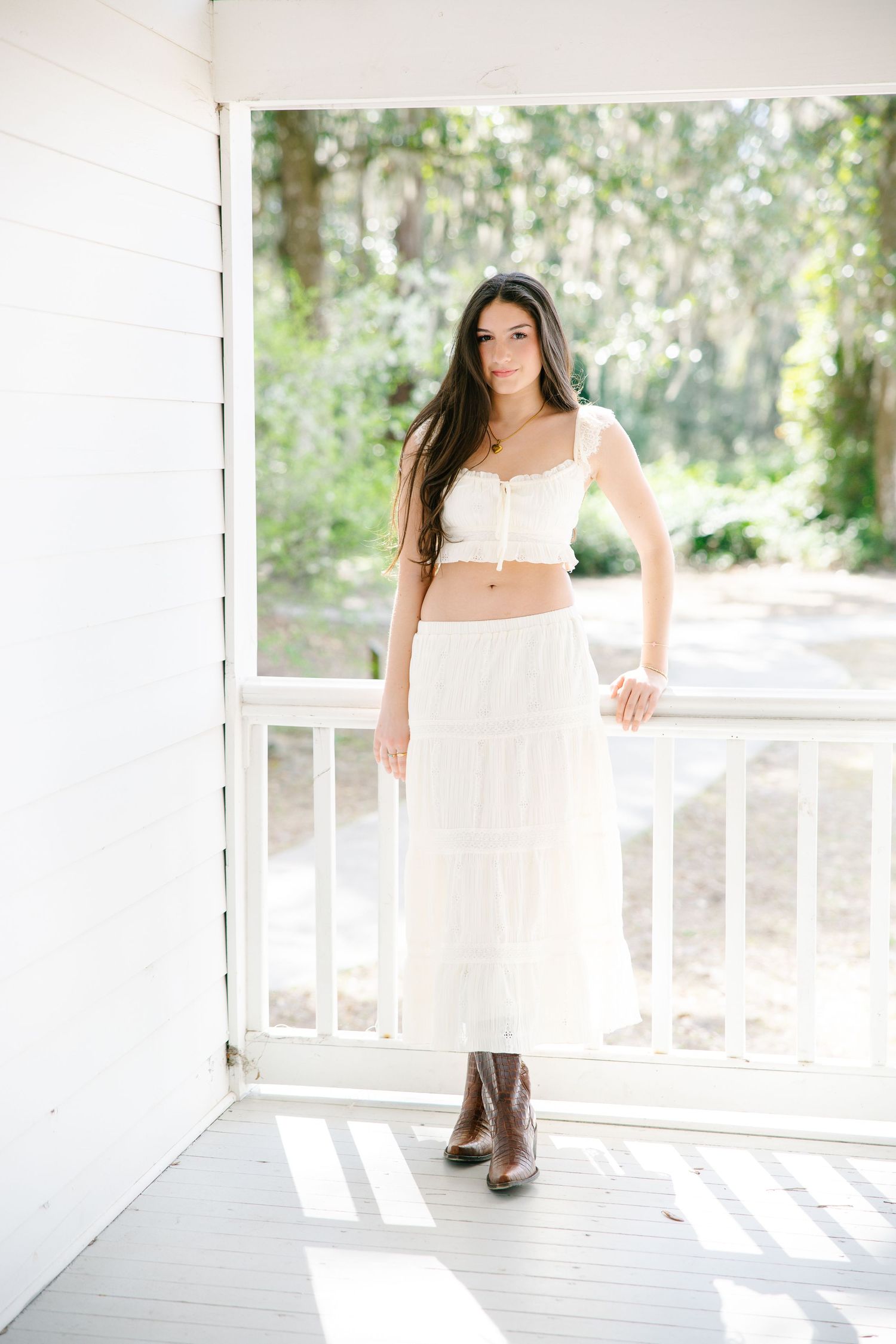 Smiling young woman in white crop top, tiered midi skirt, and brown boots on bright white porch.