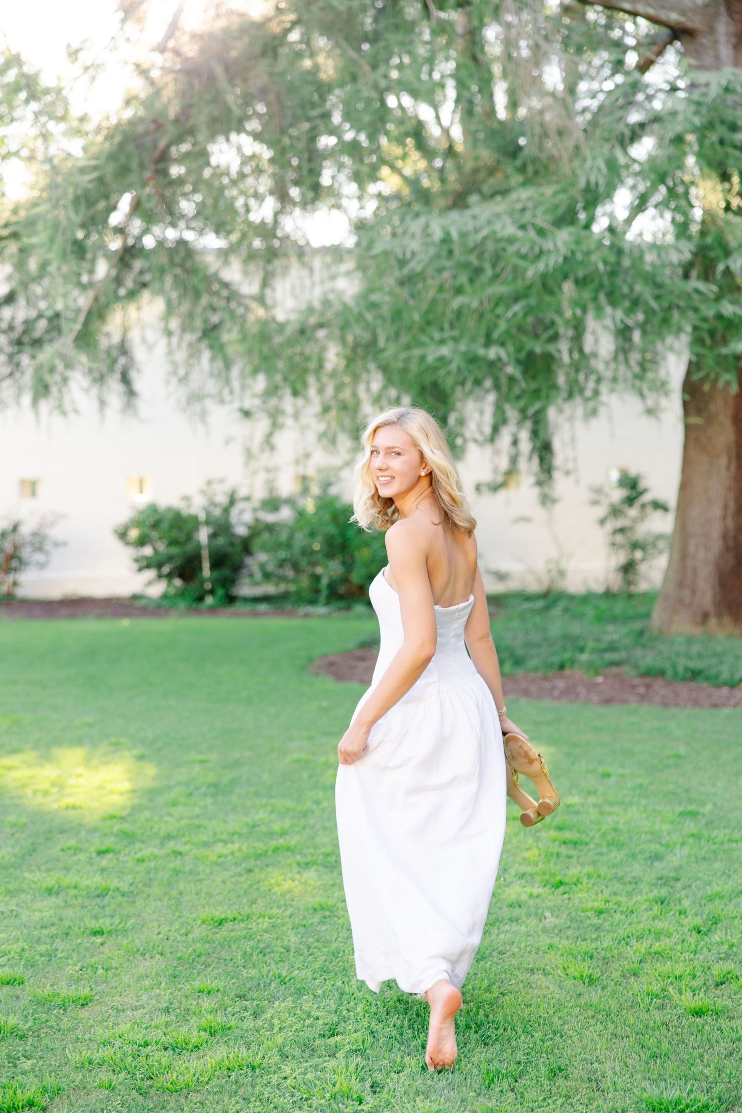 Blonde woman in white strapless dress walking barefoot on green lawn near large tree.