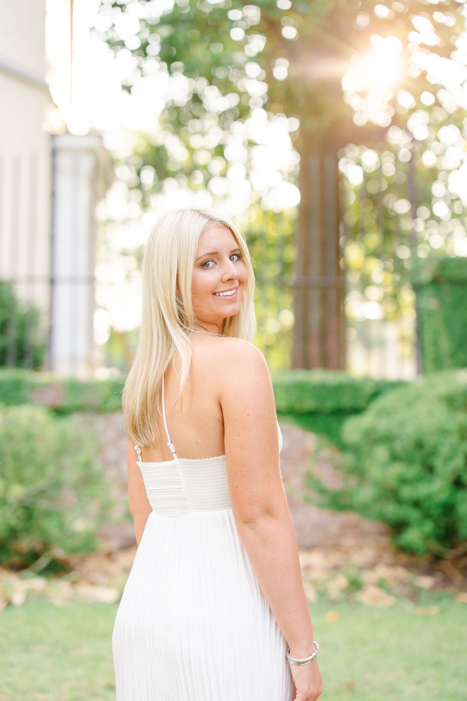 Blonde woman in white pleated spaghetti strap dress turning to smile in sunlit garden setting.