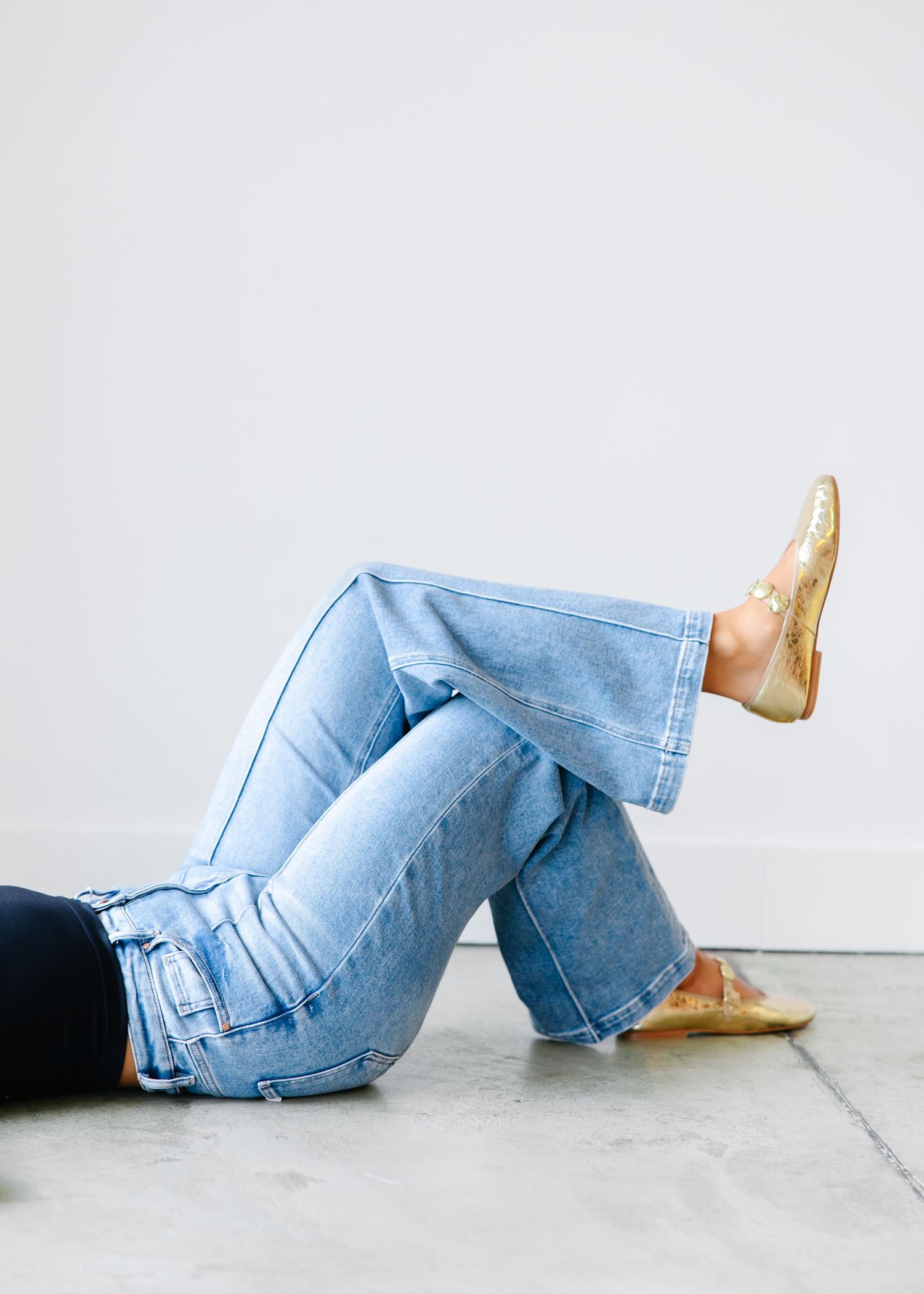 Close-up of light wash wide-leg jeans with seam detailing and gold metallic flats on a seated model.