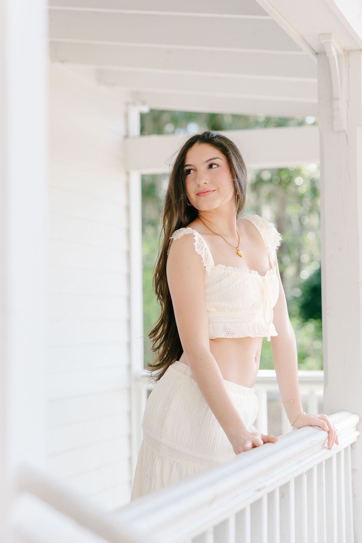 Brunette teenage girl in cream crop top and skirt set posing on white porch railing outdoors.