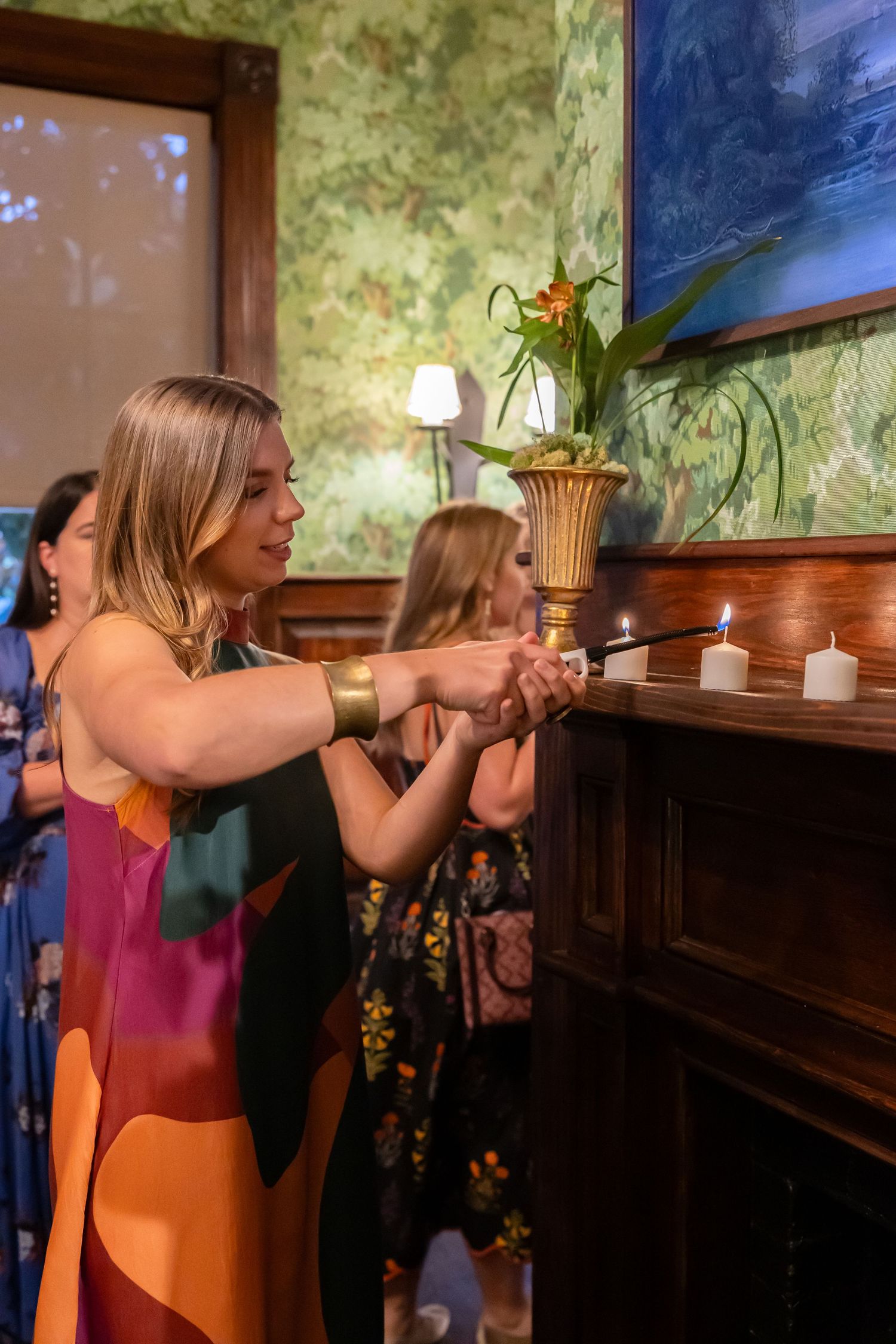 Women in colorful dresses lighting candles on a fireplace mantel decorated with flowers and green wallpaper backdrop.