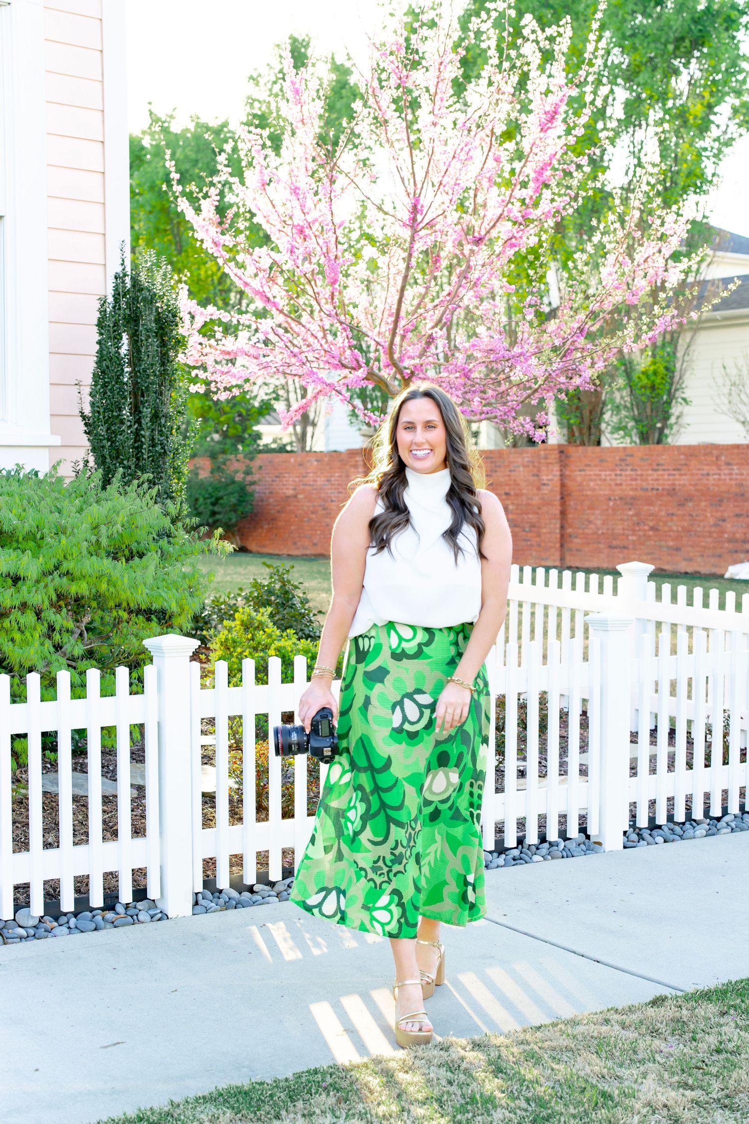 Smiling woman in white top and green floral skirt holds camera near a blooming pink tree.