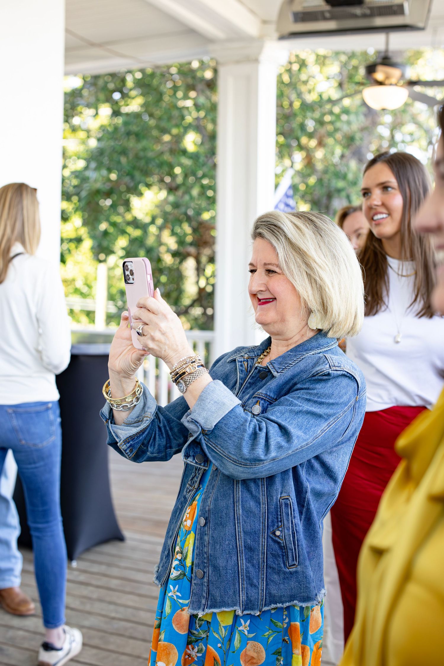 Woman in denim jacket smiling while taking a photo on her smartphone at an outdoor gathering.