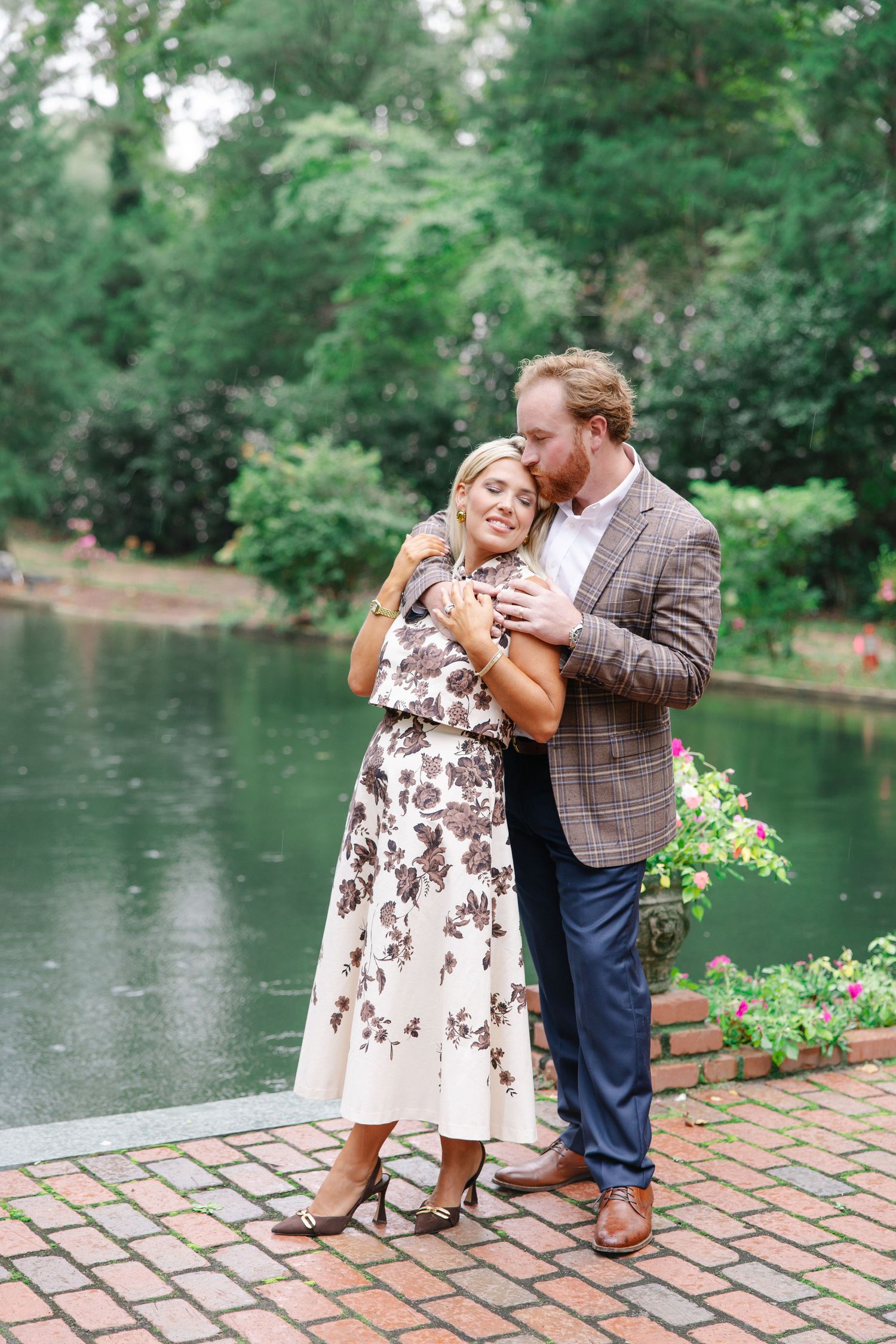 Romantic couple embracing on brick patio beside serene pond surrounded by lush greenery.