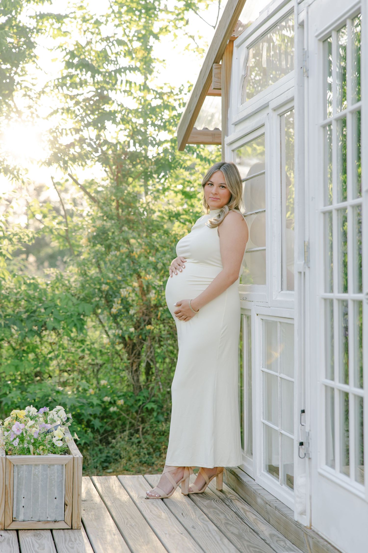 Elegant pregnant woman in white maxi dress stands on a sunlit wooden porch beside large white windows.