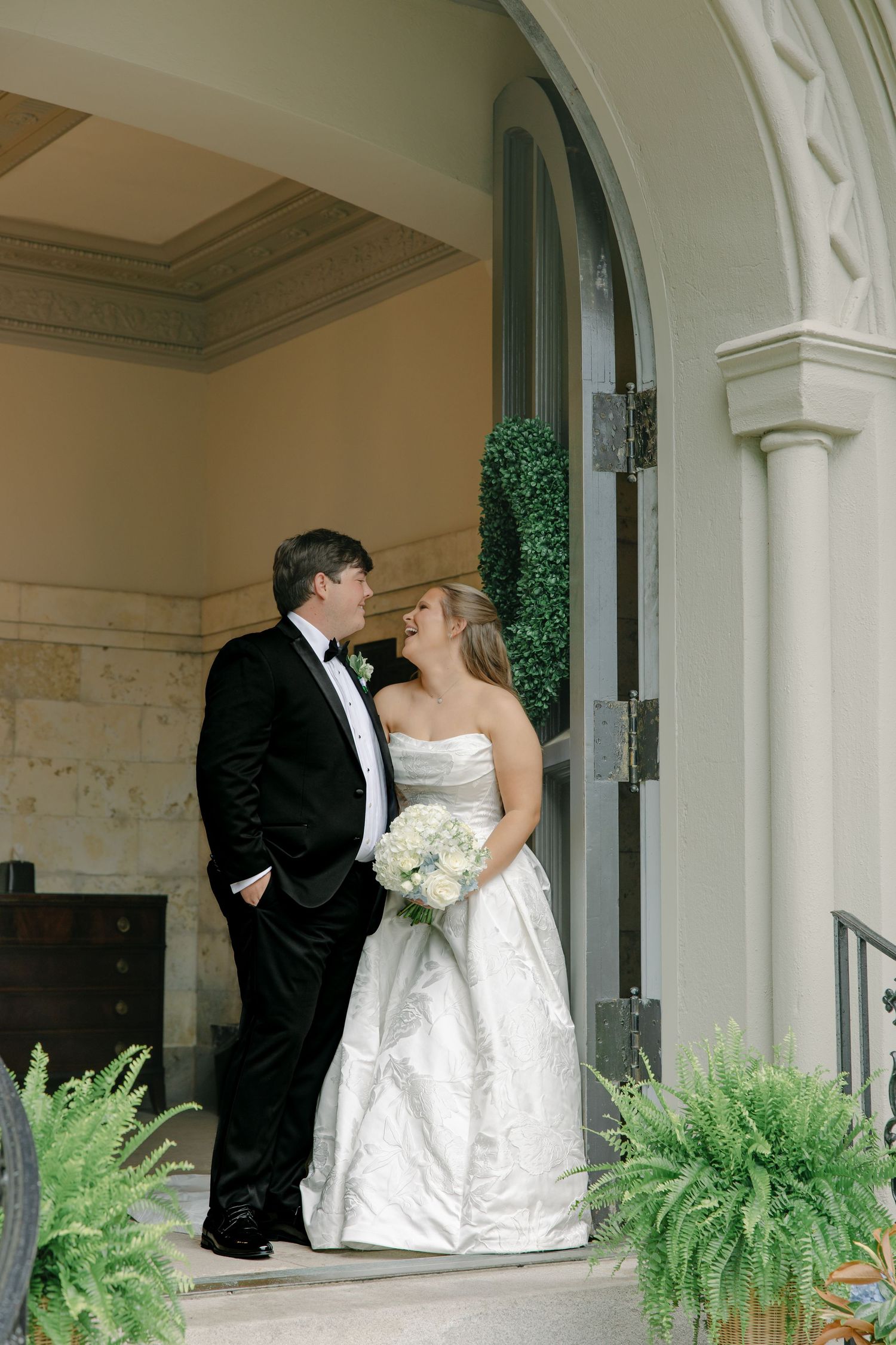 Bride and groom share romantic glance under elegant arched doorway with lush fern decor.