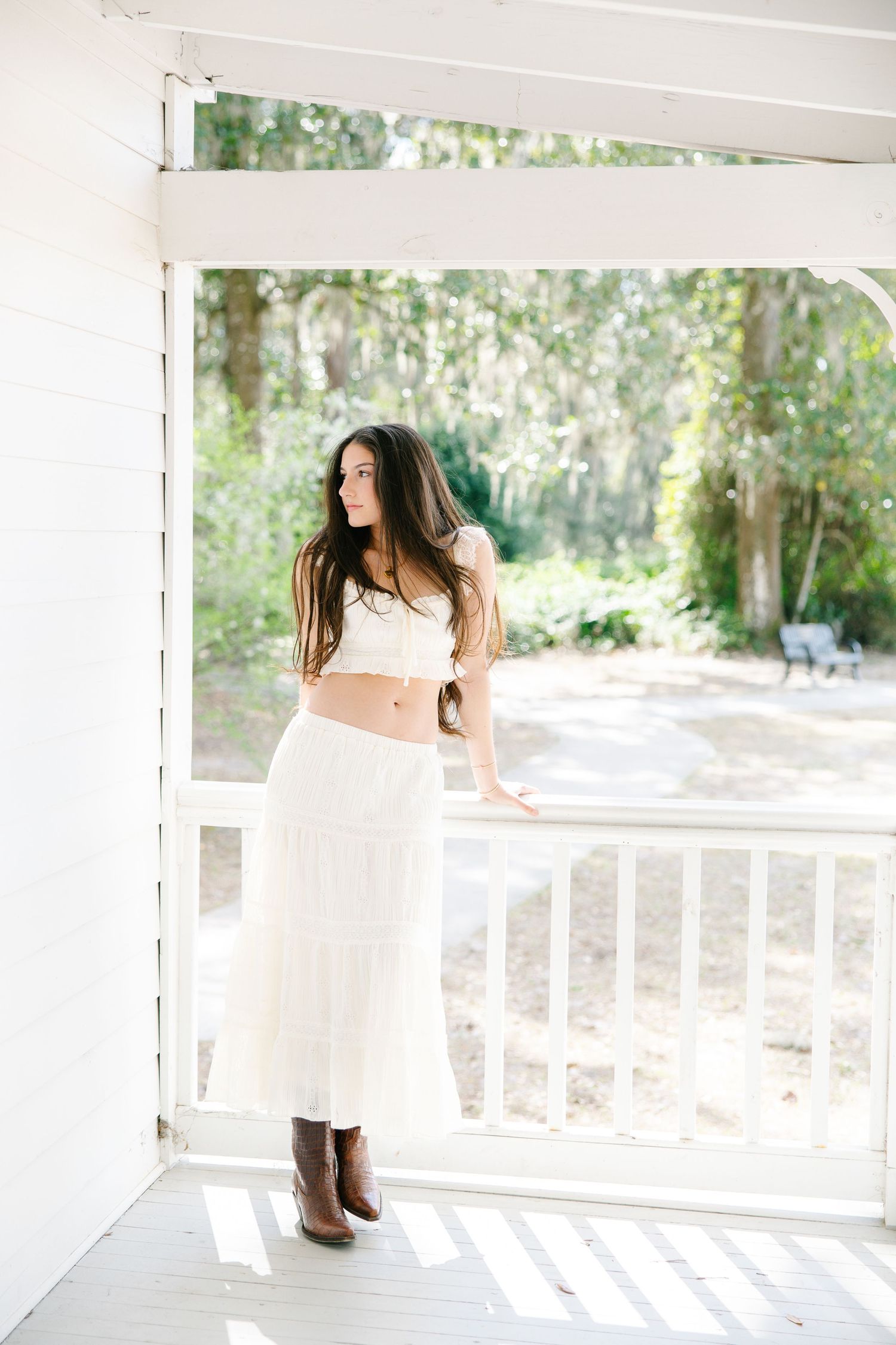 Young woman in white crop top and maxi skirt with brown boots stands on sunlit porch.