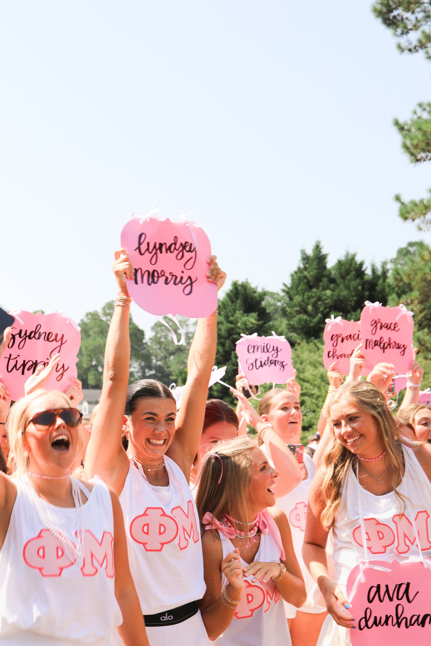 Excited Phi Mu sorority members in white tanks holding pink name signs during bid day.