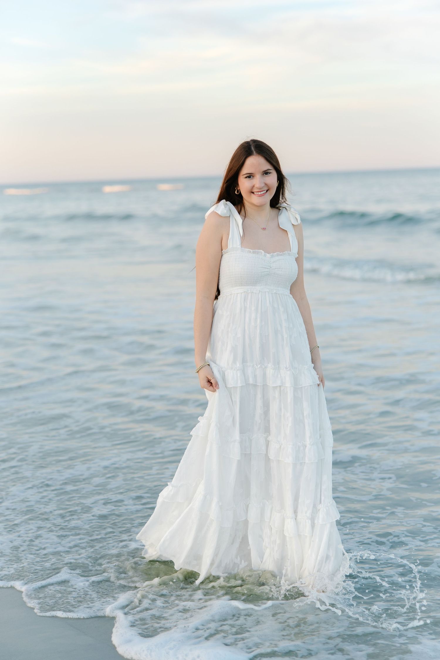 Happy young woman in white tiered maxi dress standing in ocean waves at sunset beach.