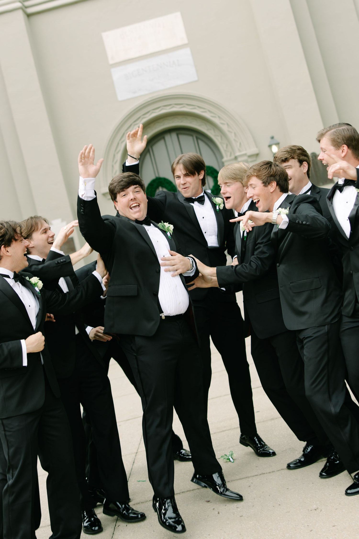 Groom celebrating joyfully with groomsmen in black tuxedos outside church on wedding day.