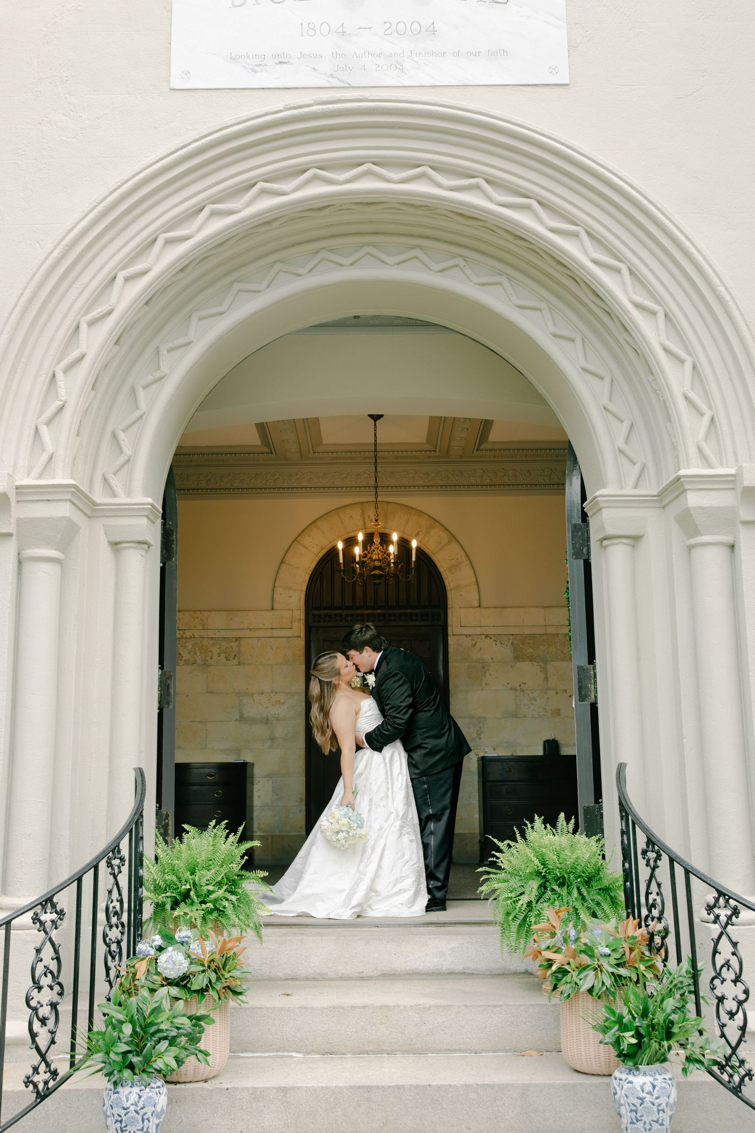 Bride and groom sharing a kiss under an ornate arched doorway of an elegant white church with lush greenery.