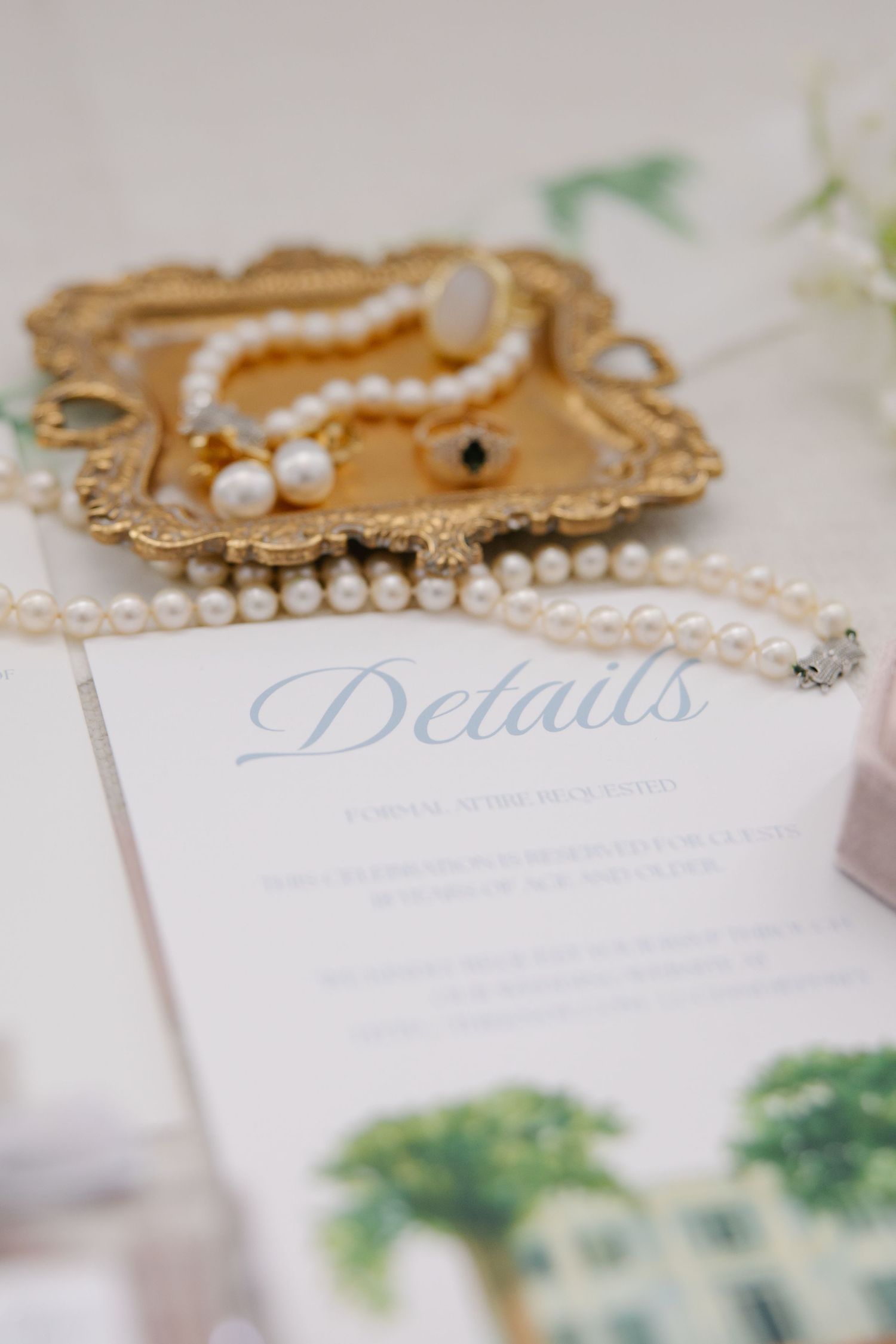 Pearl necklace and earrings on gold tray beside elegant wedding details card with watercolor venue illustration.