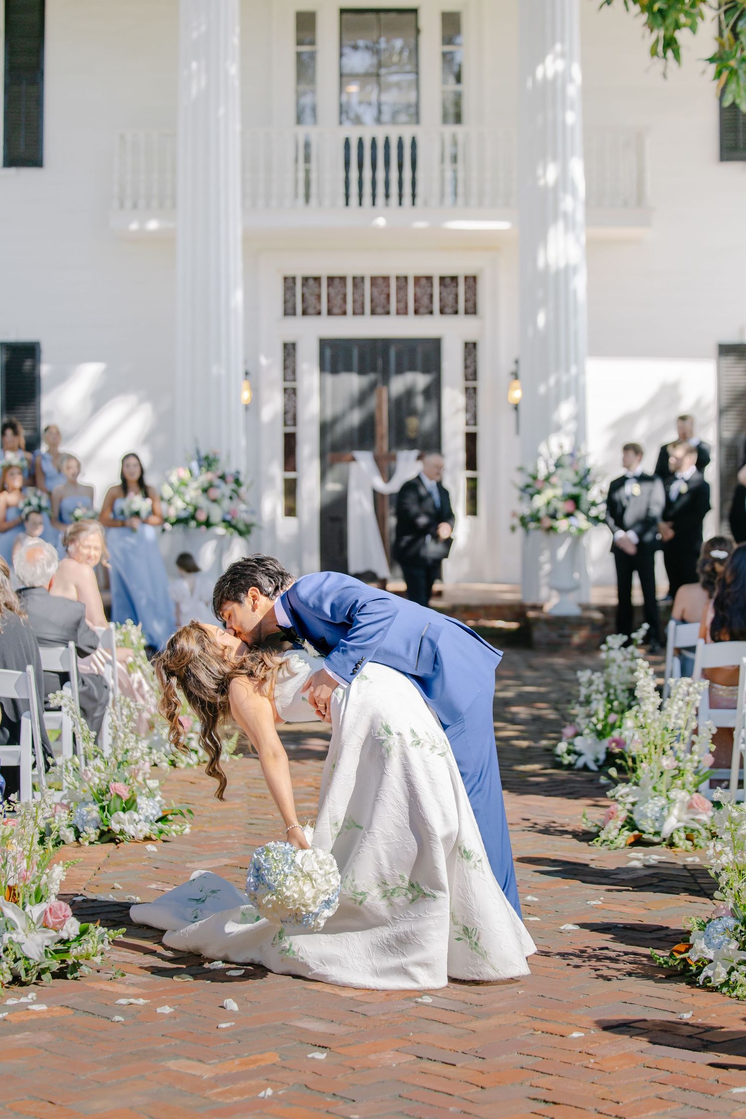 Groom in blue suit dipping bride in white gown for kiss after outdoor wedding ceremony.