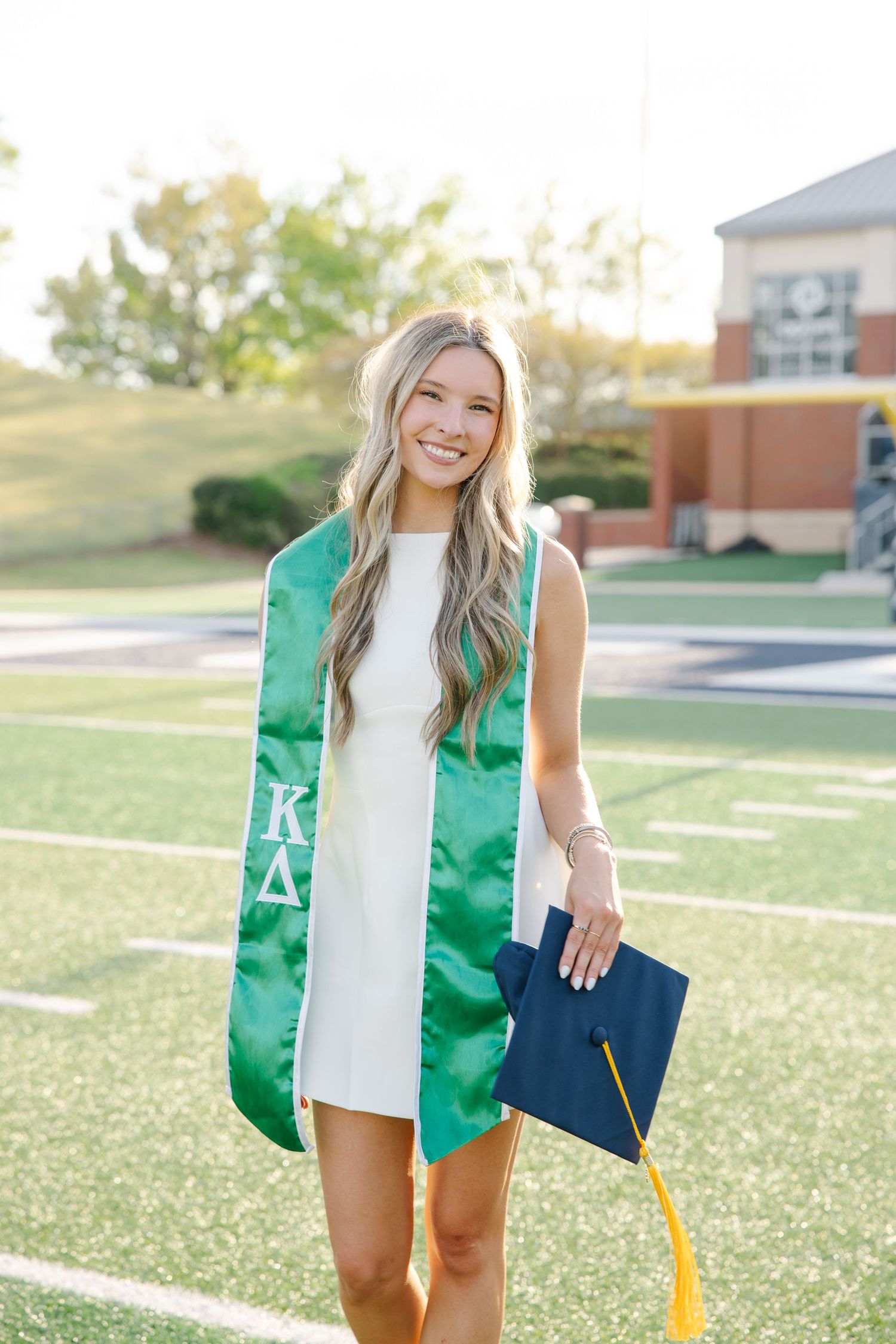 Smiling female graduate in white dress with Kappa Delta green stole holding navy graduation cap.