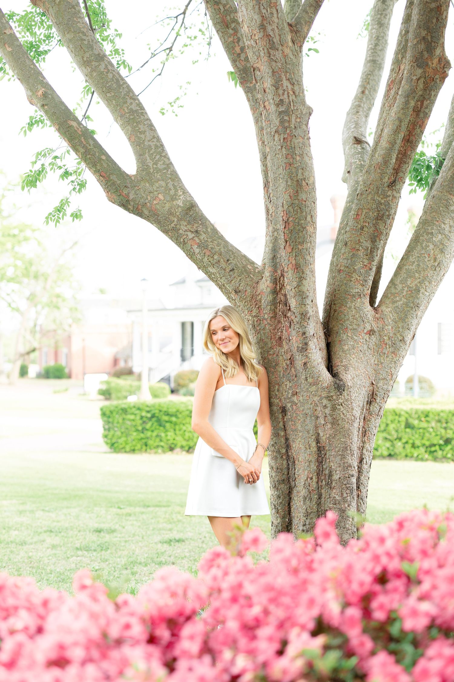 Blonde woman in white dress smiles beside large tree with pink azalea flowers in foreground.