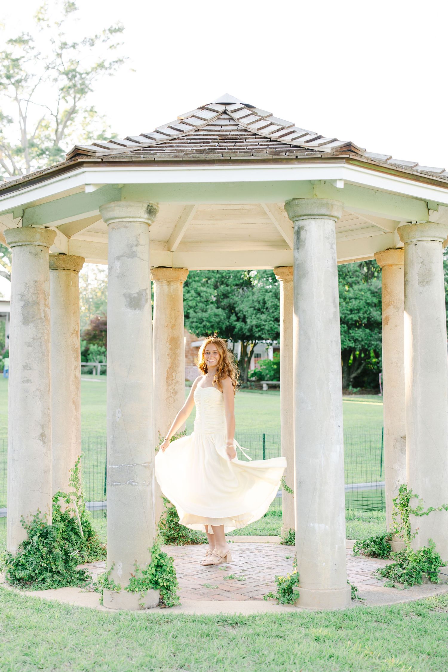 Young woman in cream dress twirling inside a white gazebo with stone columns surrounded by greenery.