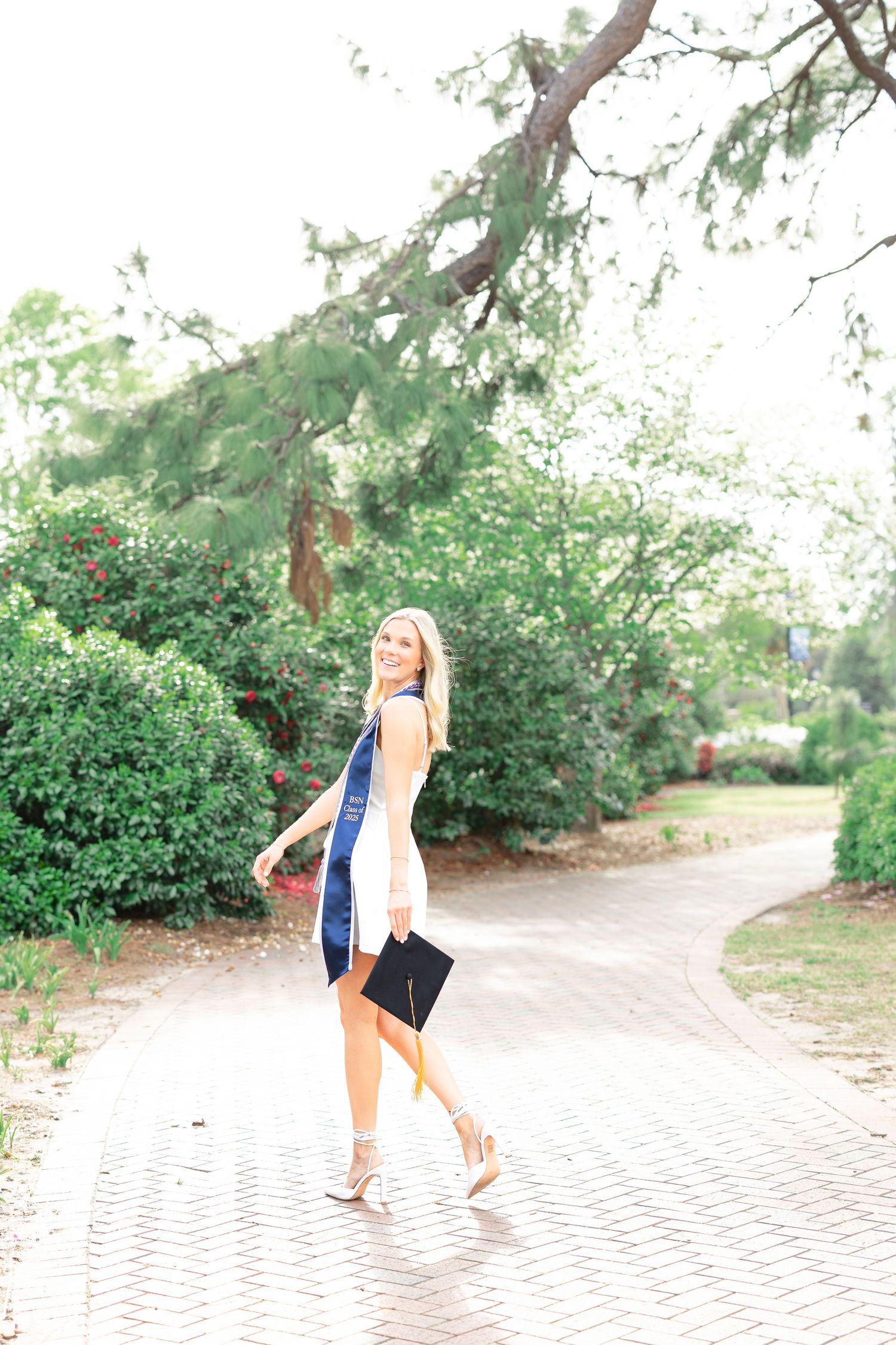 Smiling nursing graduate walks brick path in white dress with navy graduation stole holding cap.