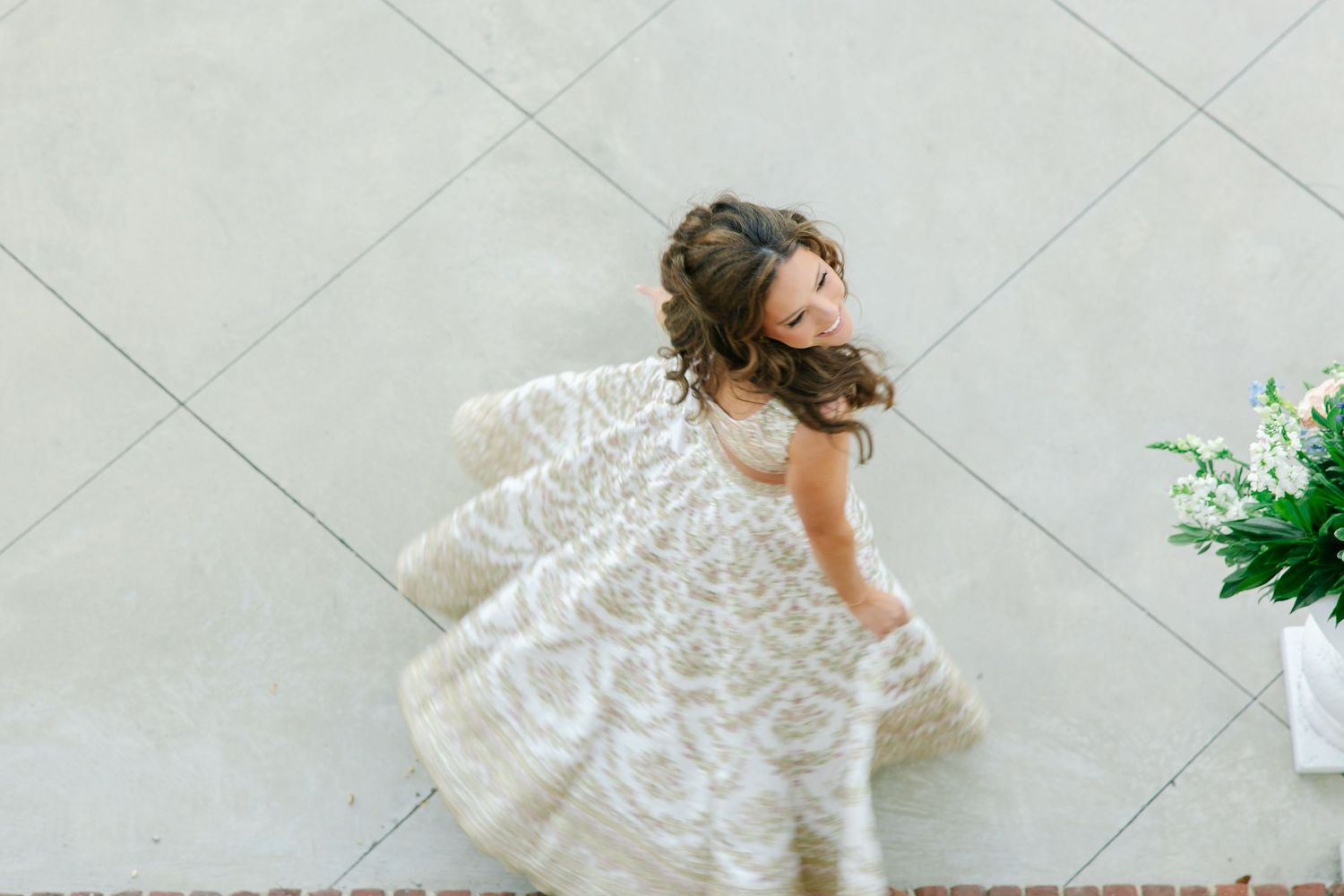 Young flower girl twirls in elegant lace dress, captured from above against a neutral wall backdrop.
