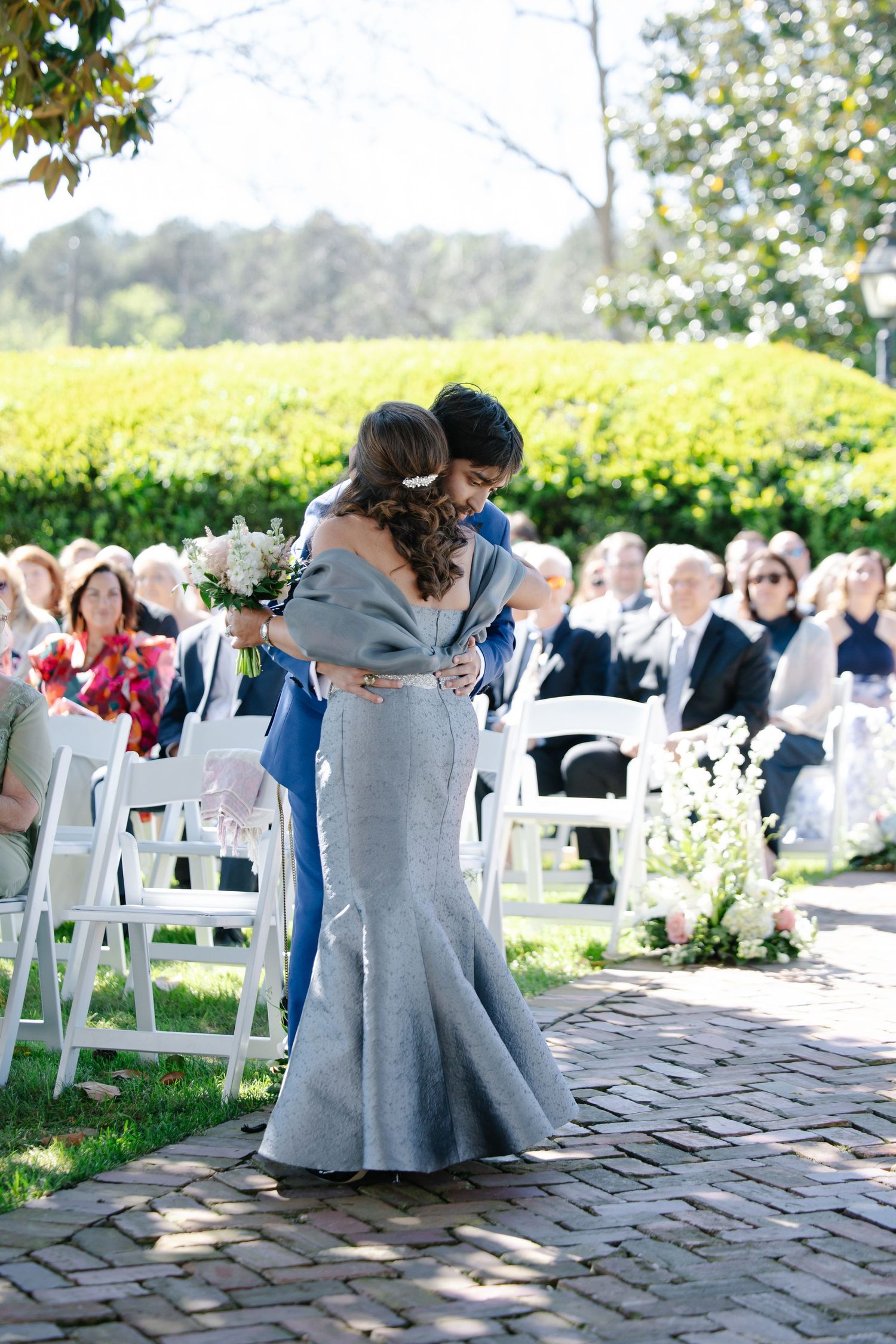 Woman in gray mermaid gown embracing groom in blue suit during outdoor wedding ceremony procession.
