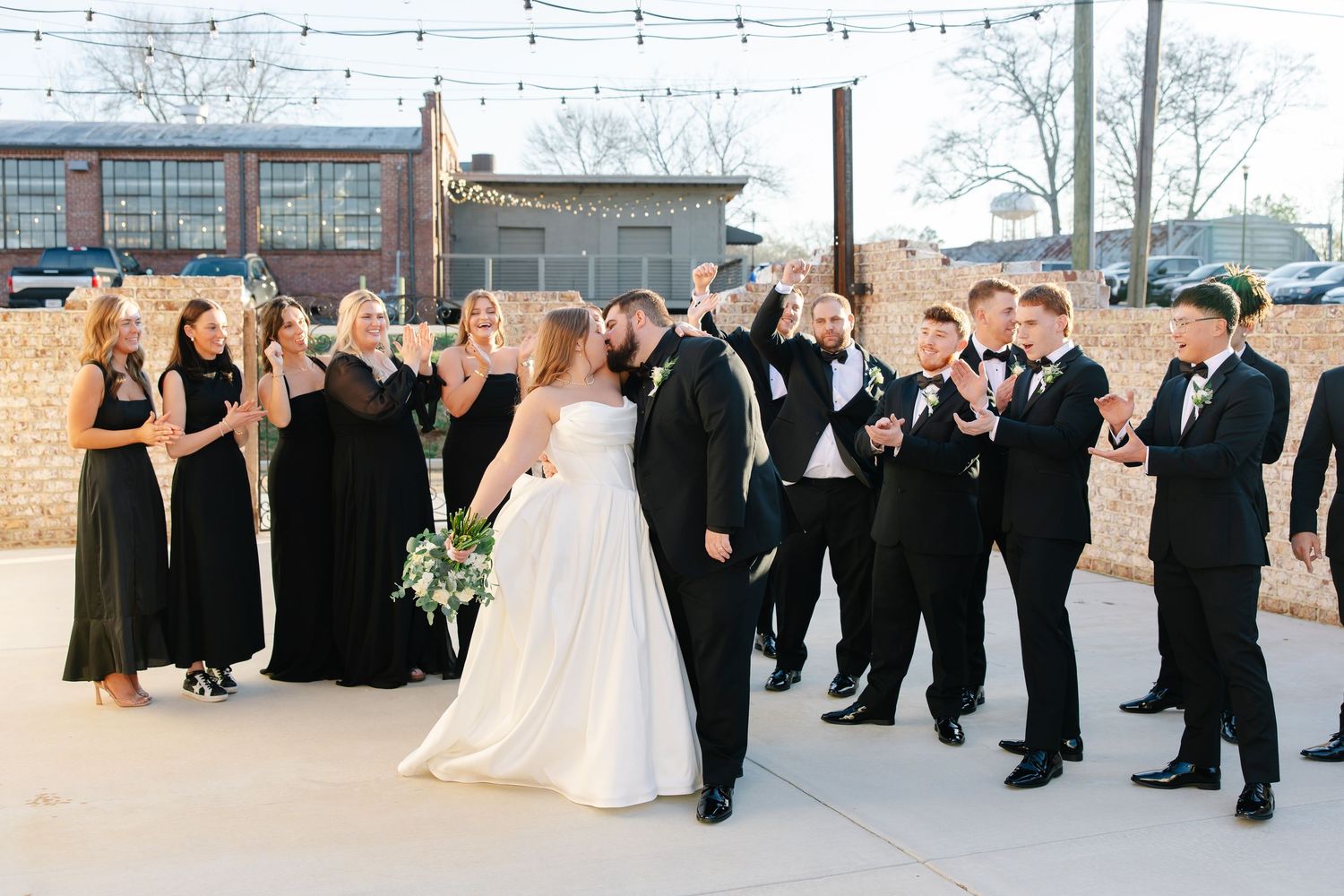Wedding party in black attire celebrates outdoors with bride in white gown, bridesmaids and groomsmen cheering together.