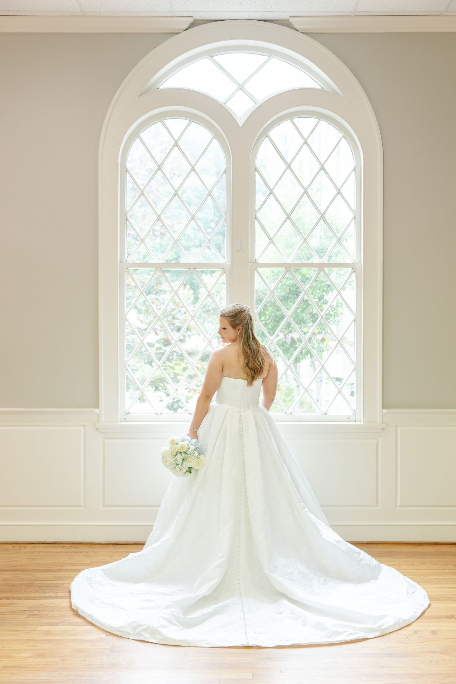 Bride in strapless white ball gown with long train poses before large arched leaded glass windows.