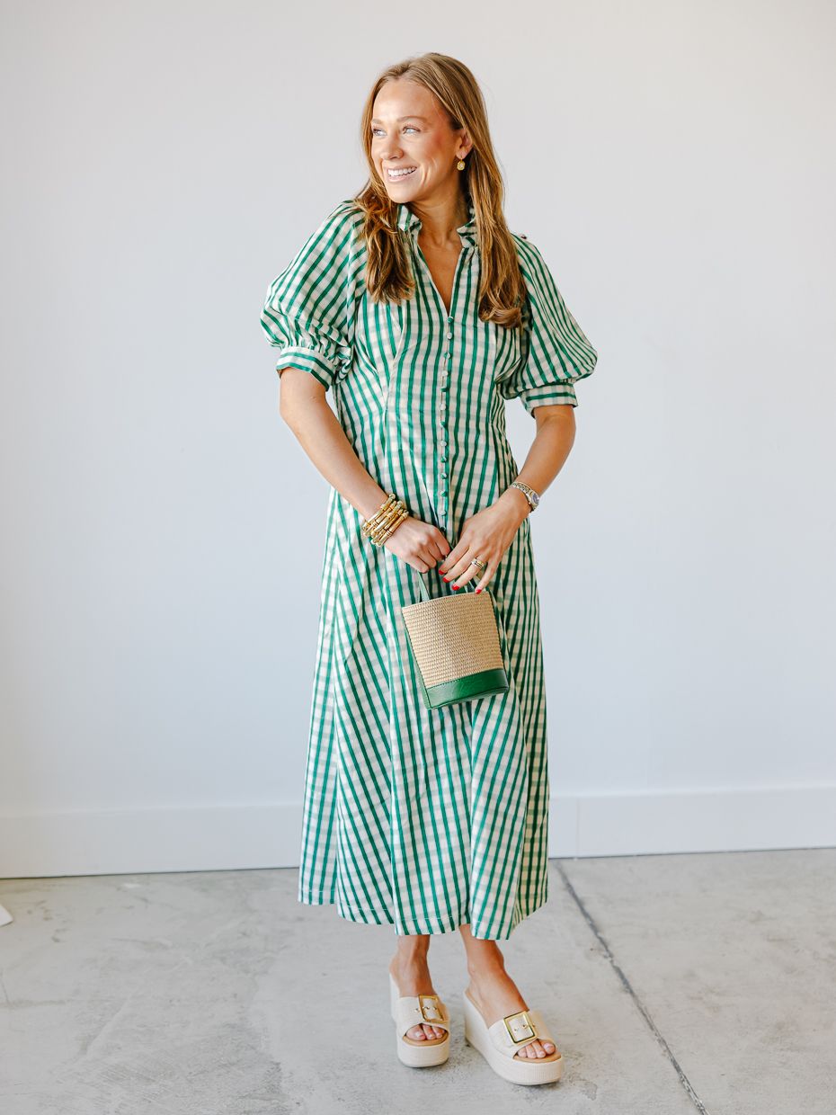 Woman wearing a green gingham midi shirtdress with puff sleeves holding a small woven bag with white sandals.