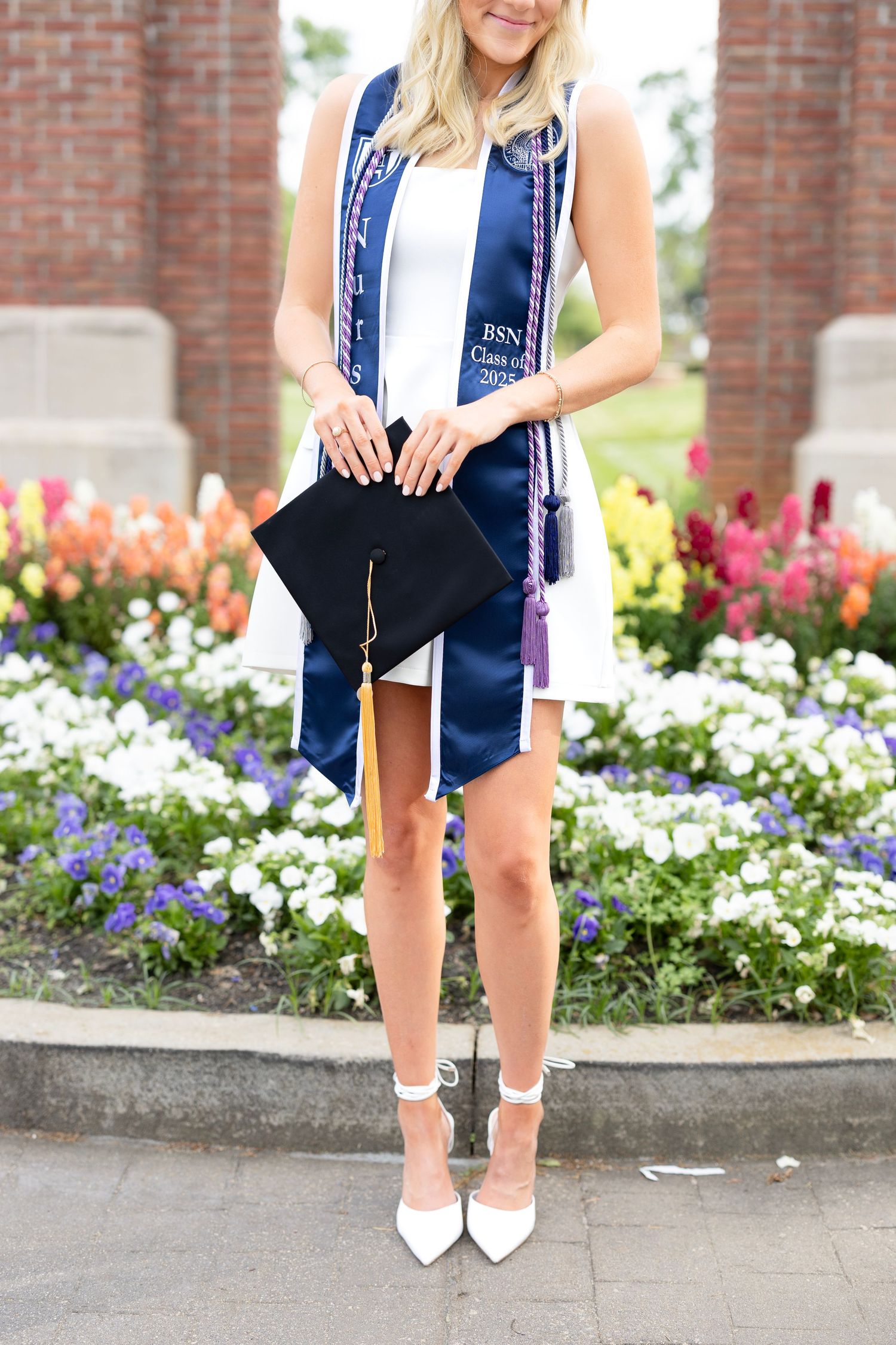 Nursing graduate in white dress with navy BSN graduation stole holds cap near colorful flower garden.