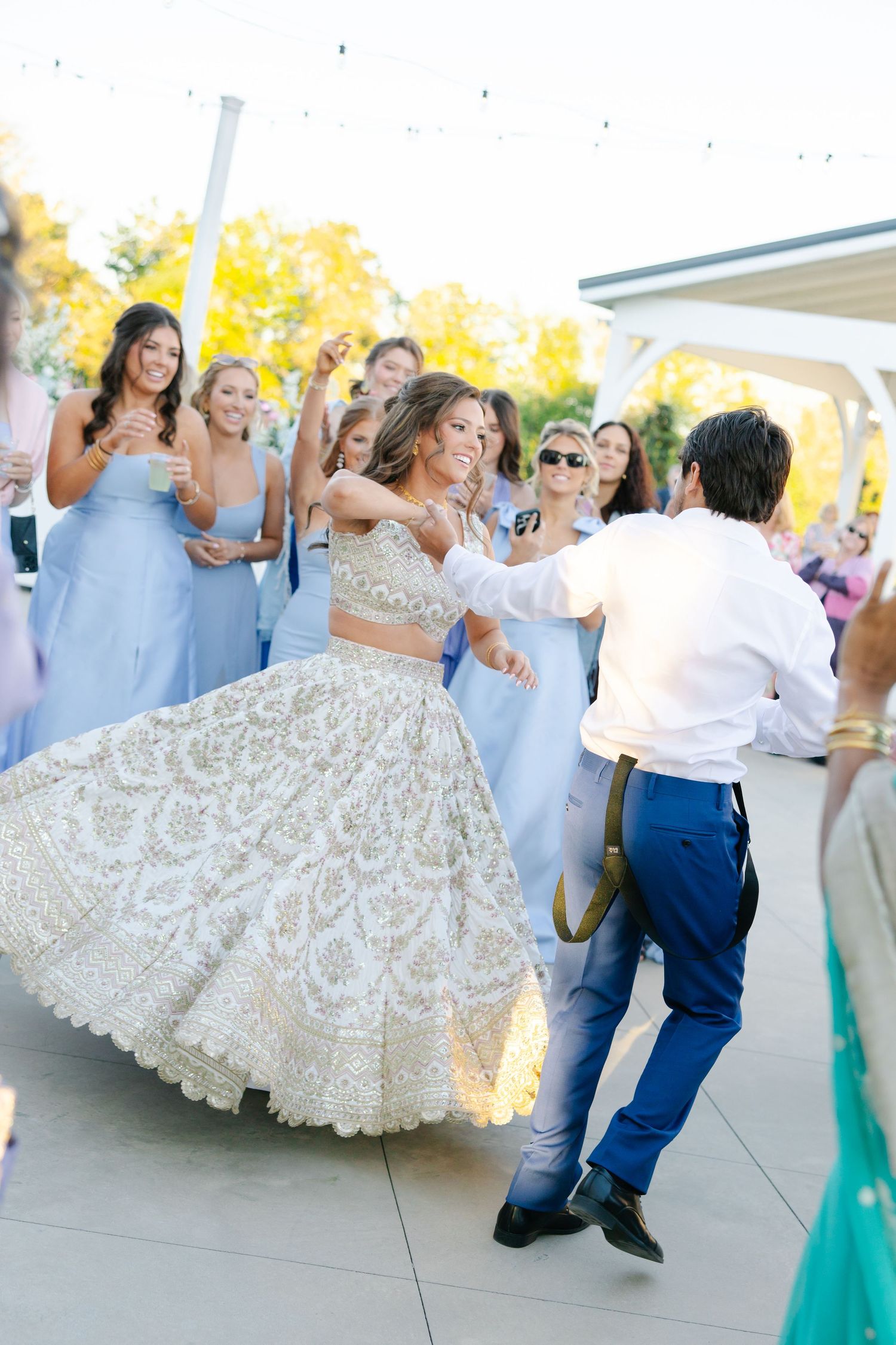 Bride in gold embroidered lehenga dancing with groom surrounded by guests at outdoor reception.