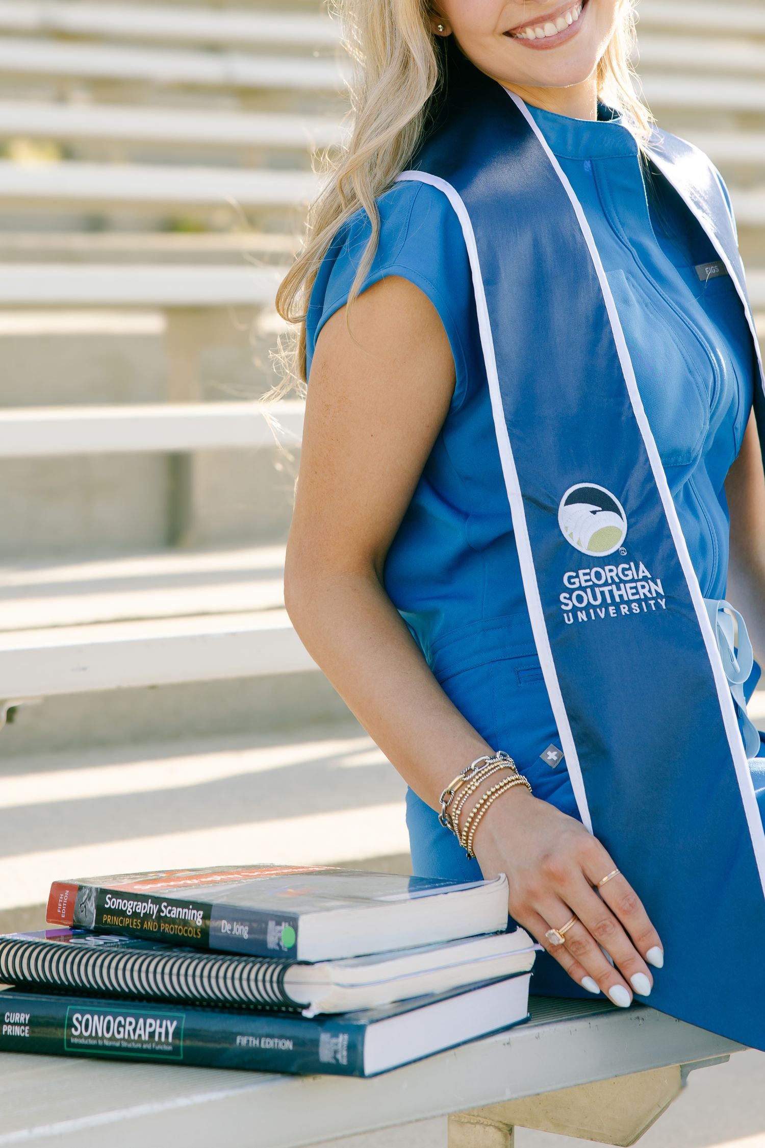 Georgia Southern University graduate in blue scrubs displaying sonography textbooks with graduation stole.