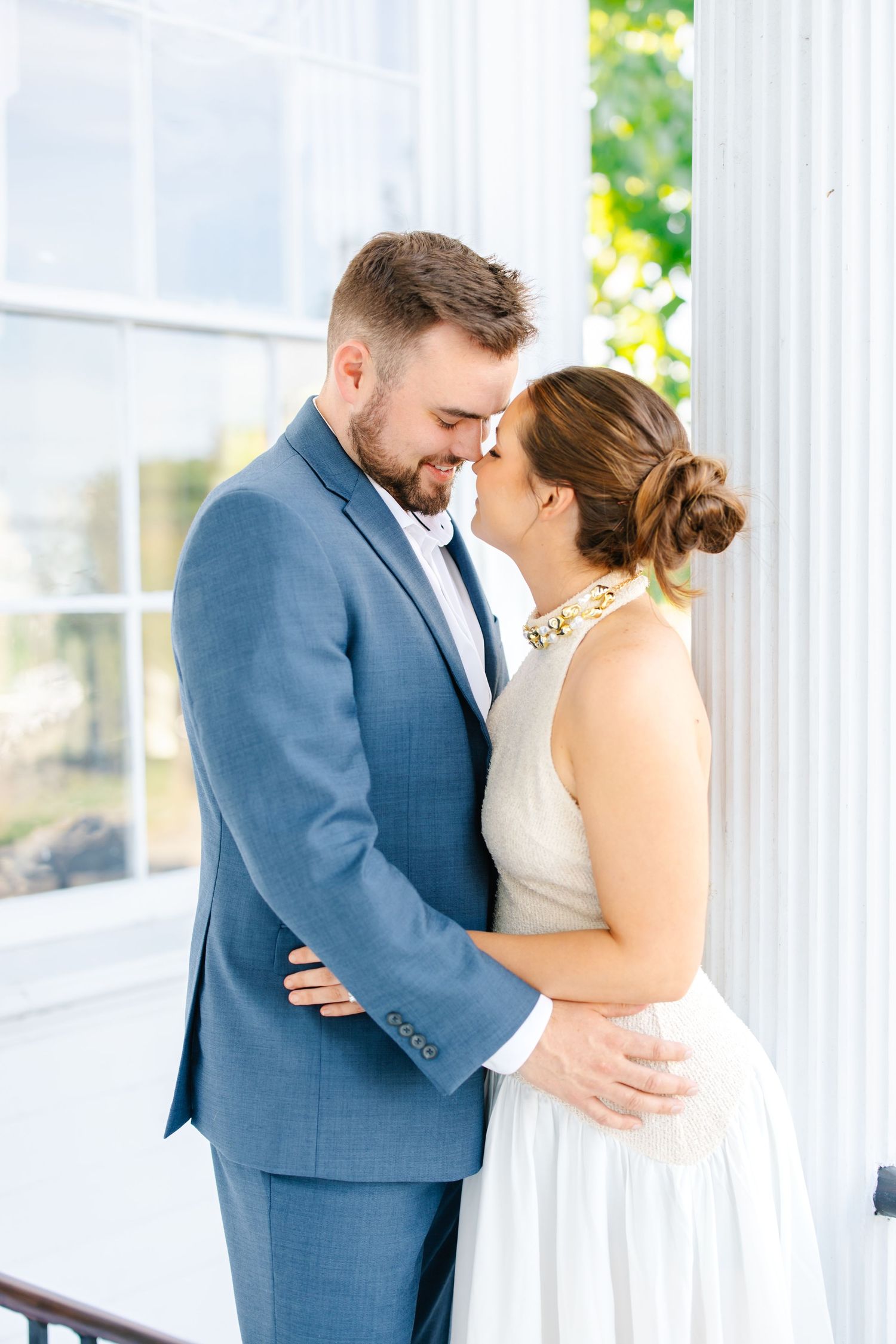 Bride and groom in tender embrace near white columns, groom in blue suit and bride in sleeveless white dress.