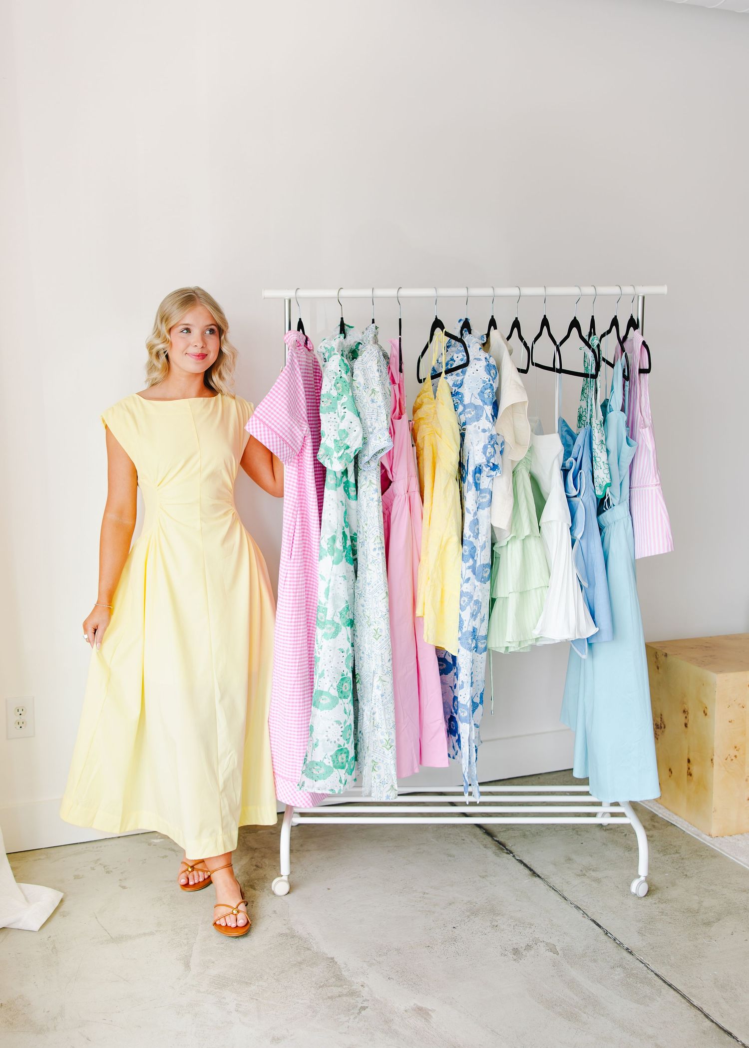 Woman in yellow midi dress stands beside a clothing rack of pastel spring dresses in pink, green, and blue.