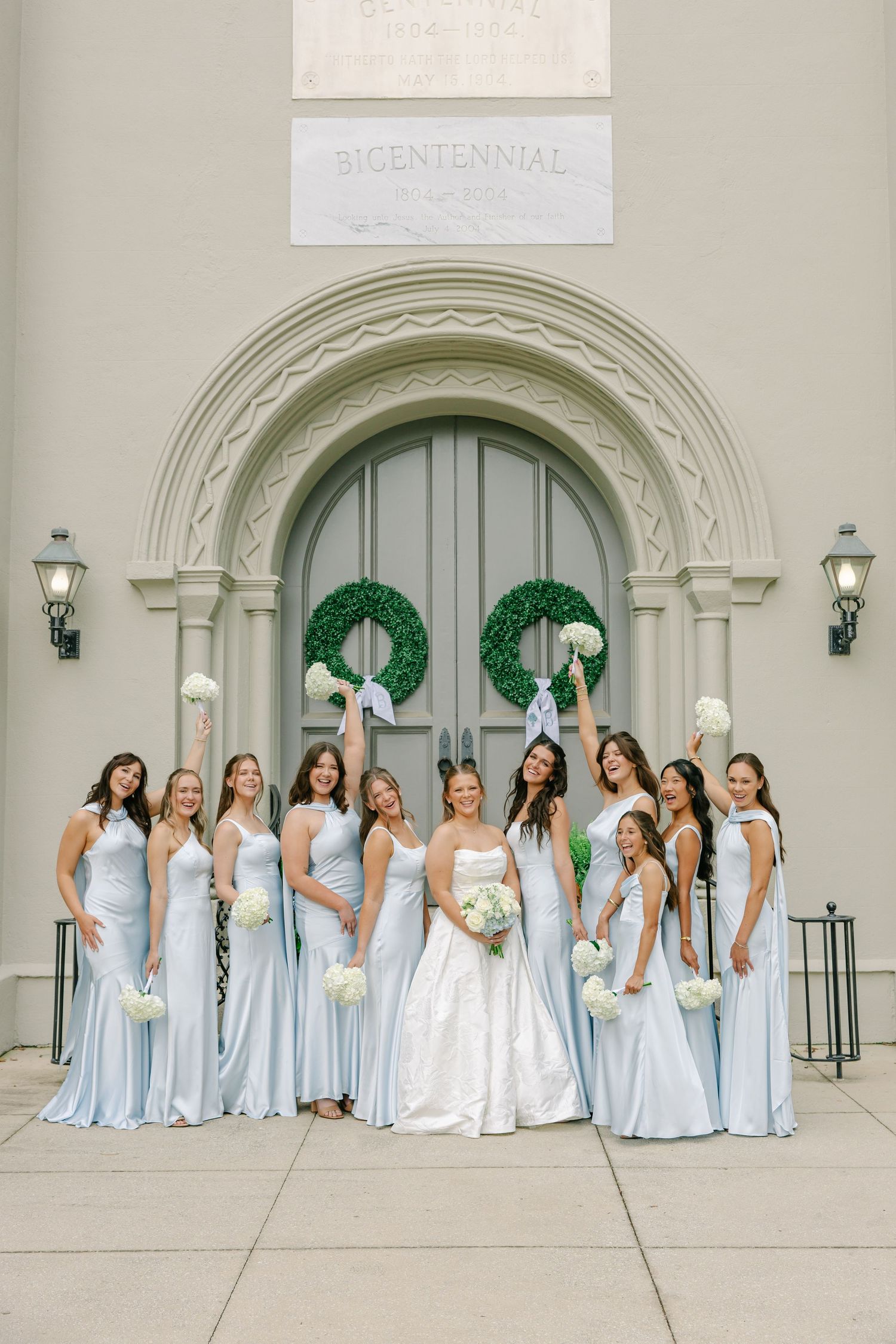 Bride and bridesmaids in light blue gowns pose joyfully before an arched church entrance with green wreaths.