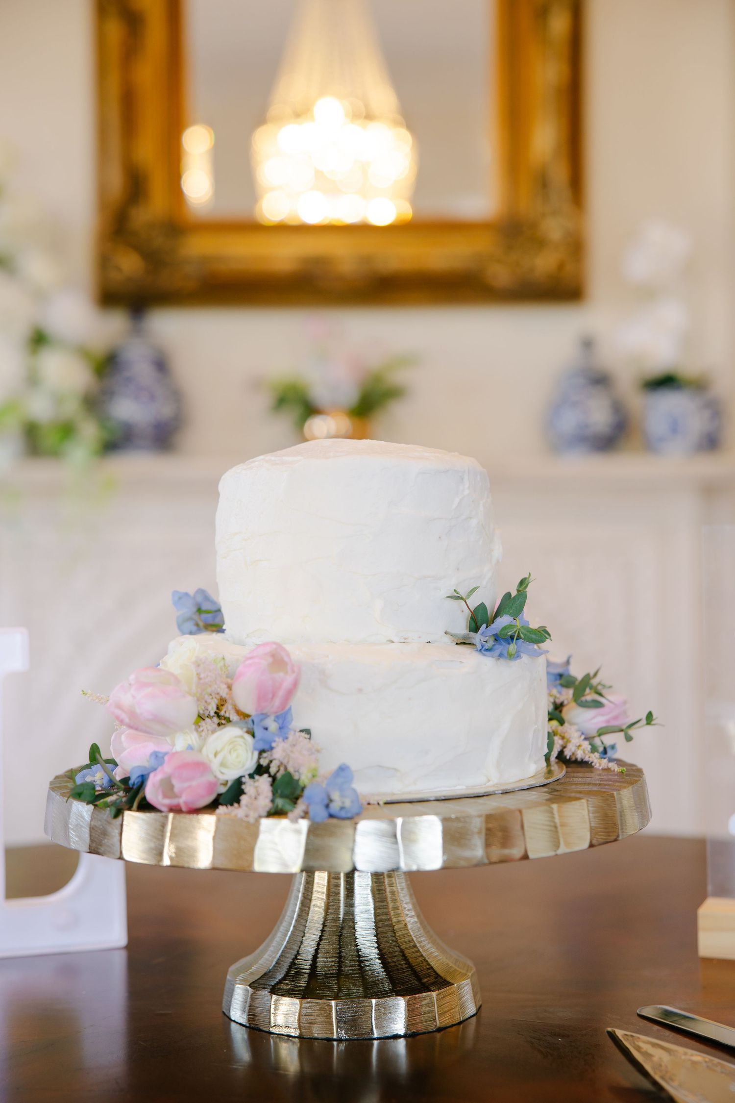 Elegant two-tier white wedding cake with pink and blue florals on gold cake stand.