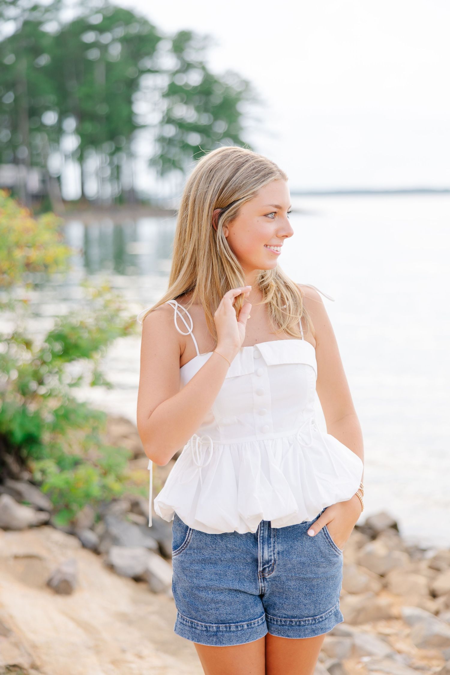 Blonde teen in white ruffled top and denim shorts standing on rocky lakeside shore looking away.