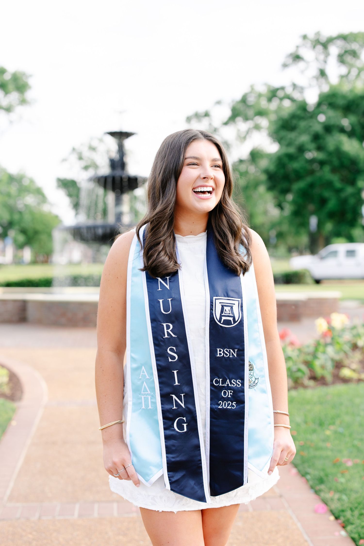 Joyful nursing BSN Class of 2025 graduate laughing in navy stole near campus fountain.