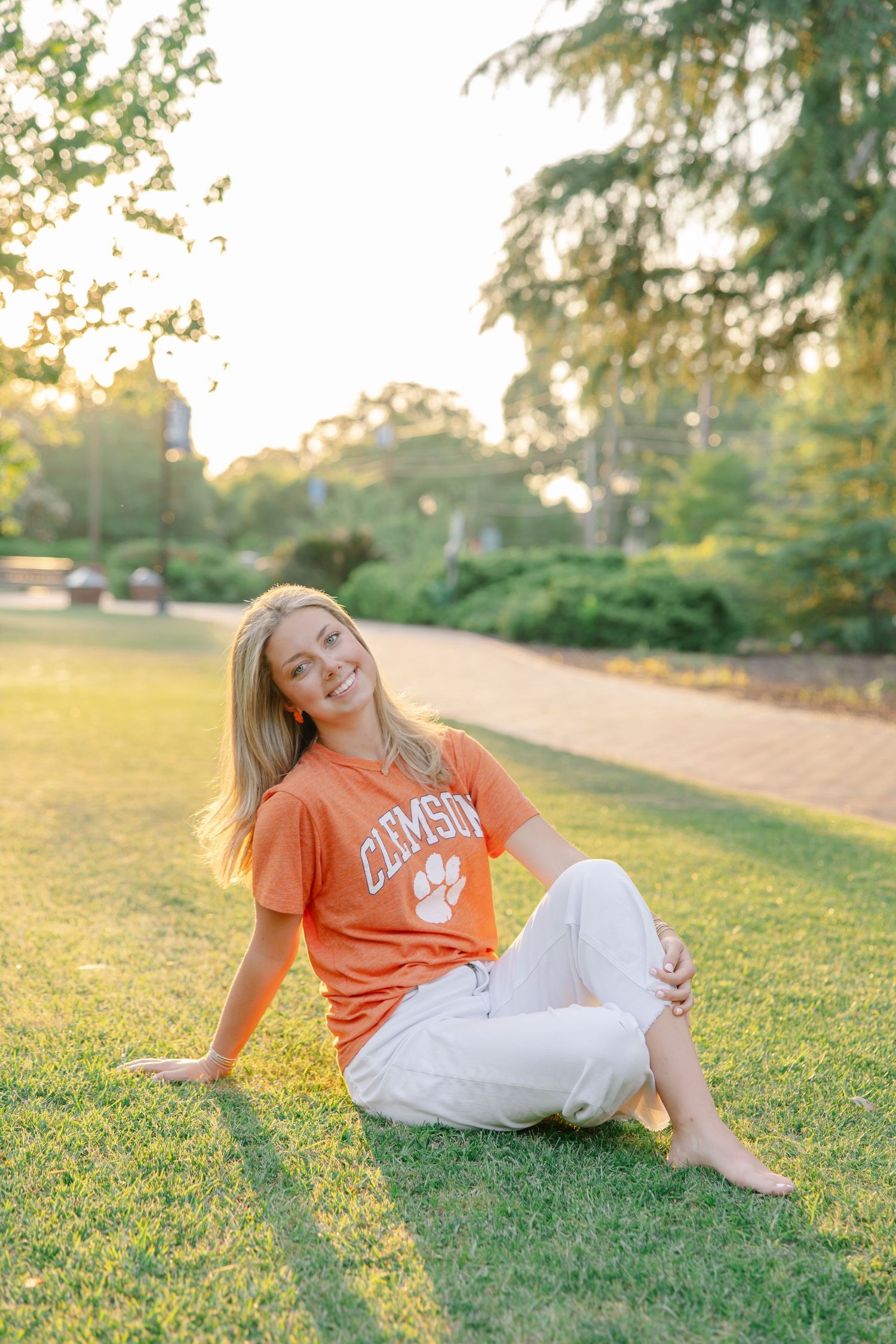 Smiling blonde woman relaxes on green grass at golden hour wearing an orange Clemson University t-shirt.
