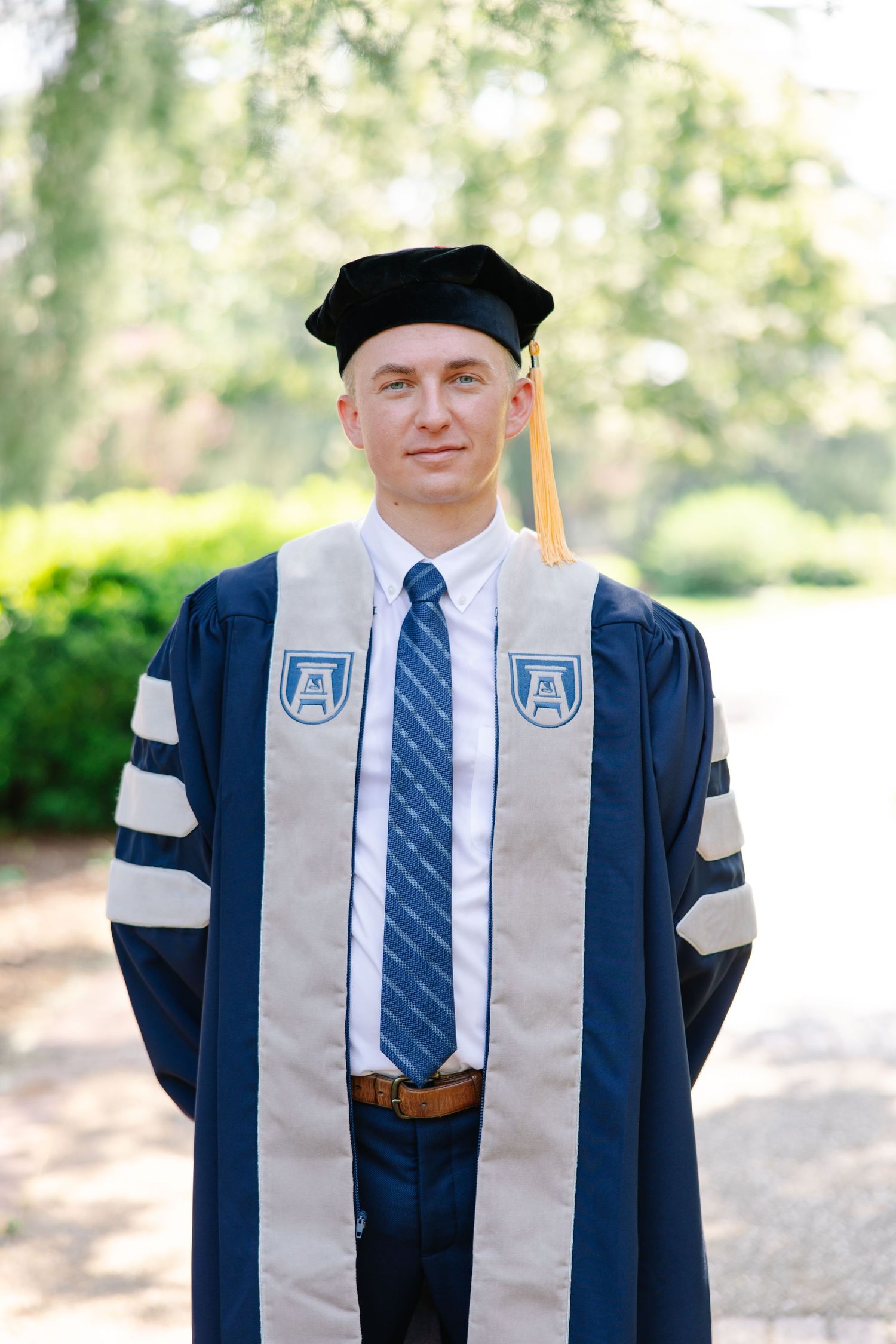 Young man in doctoral graduation regalia with navy cap and gown standing outdoors.
