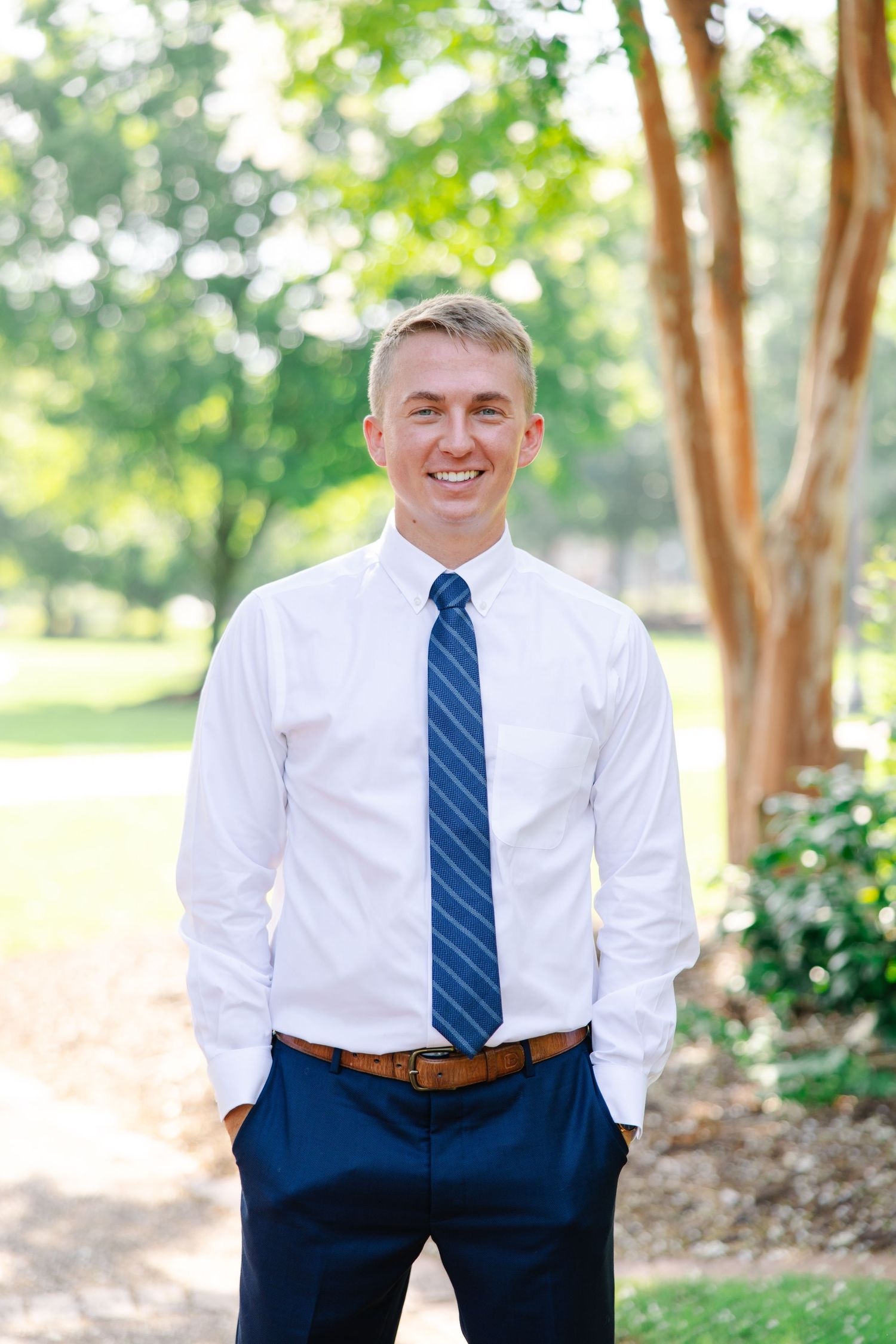 Cheerful young man in white shirt and navy tie poses outdoors among lush green trees.