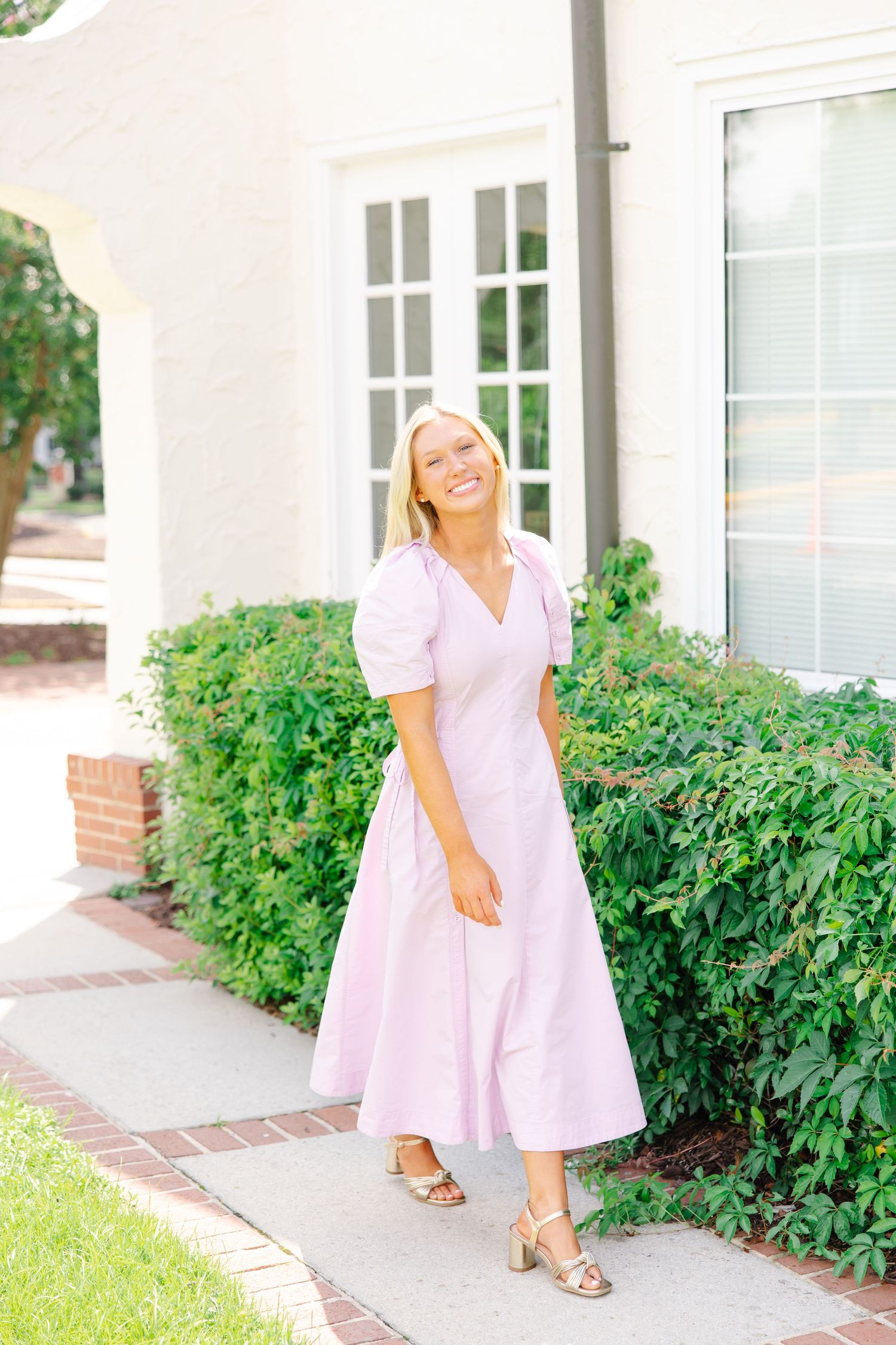 Blonde woman in lavender midi dress smiling while walking past white building and green shrubs.
